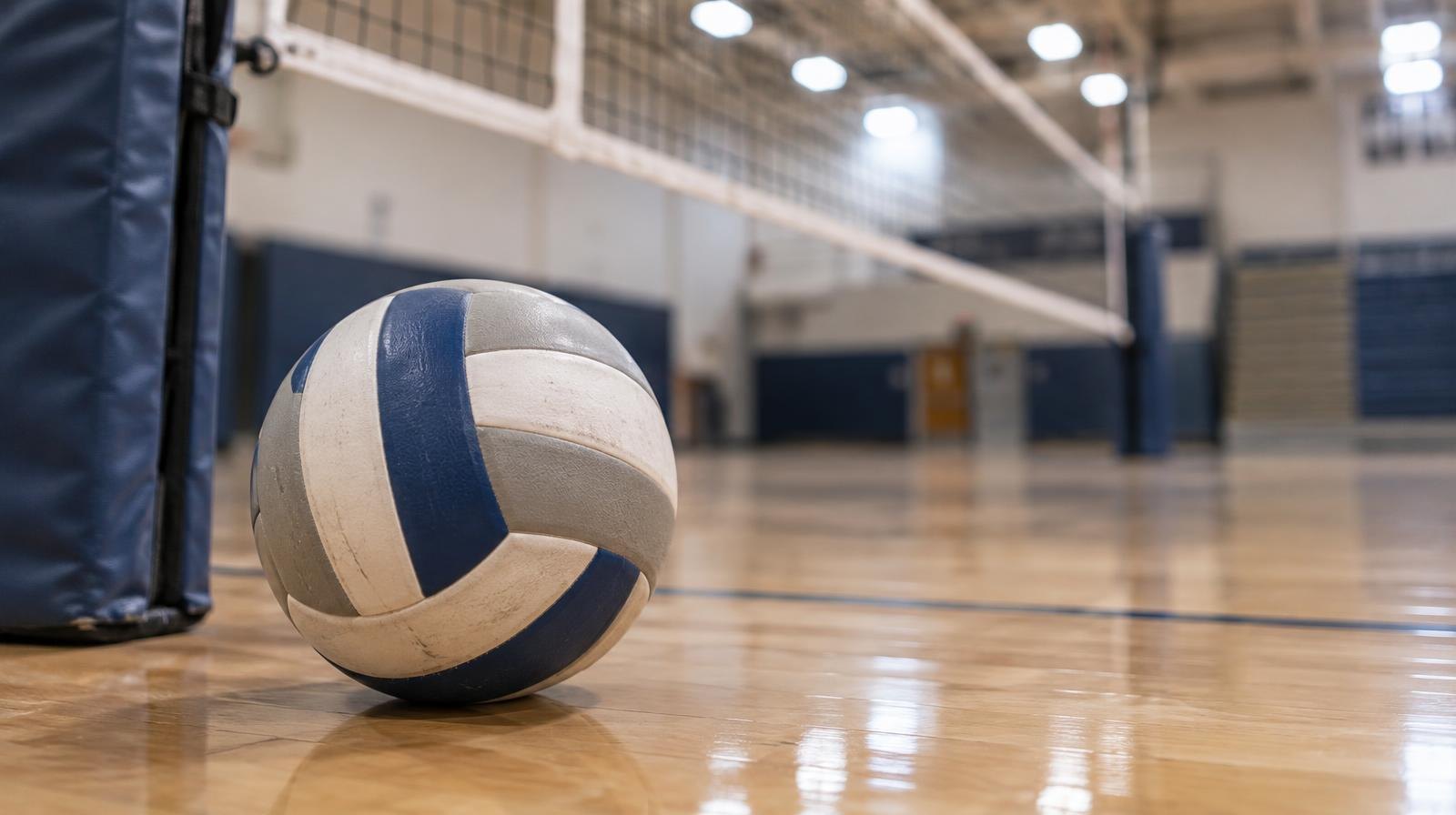 A volleyball resting on a hardwood gymnasium floor near a net post.