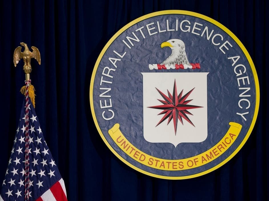 The CIA seal mounted on a blue draped background with an American flag visible to the left.
