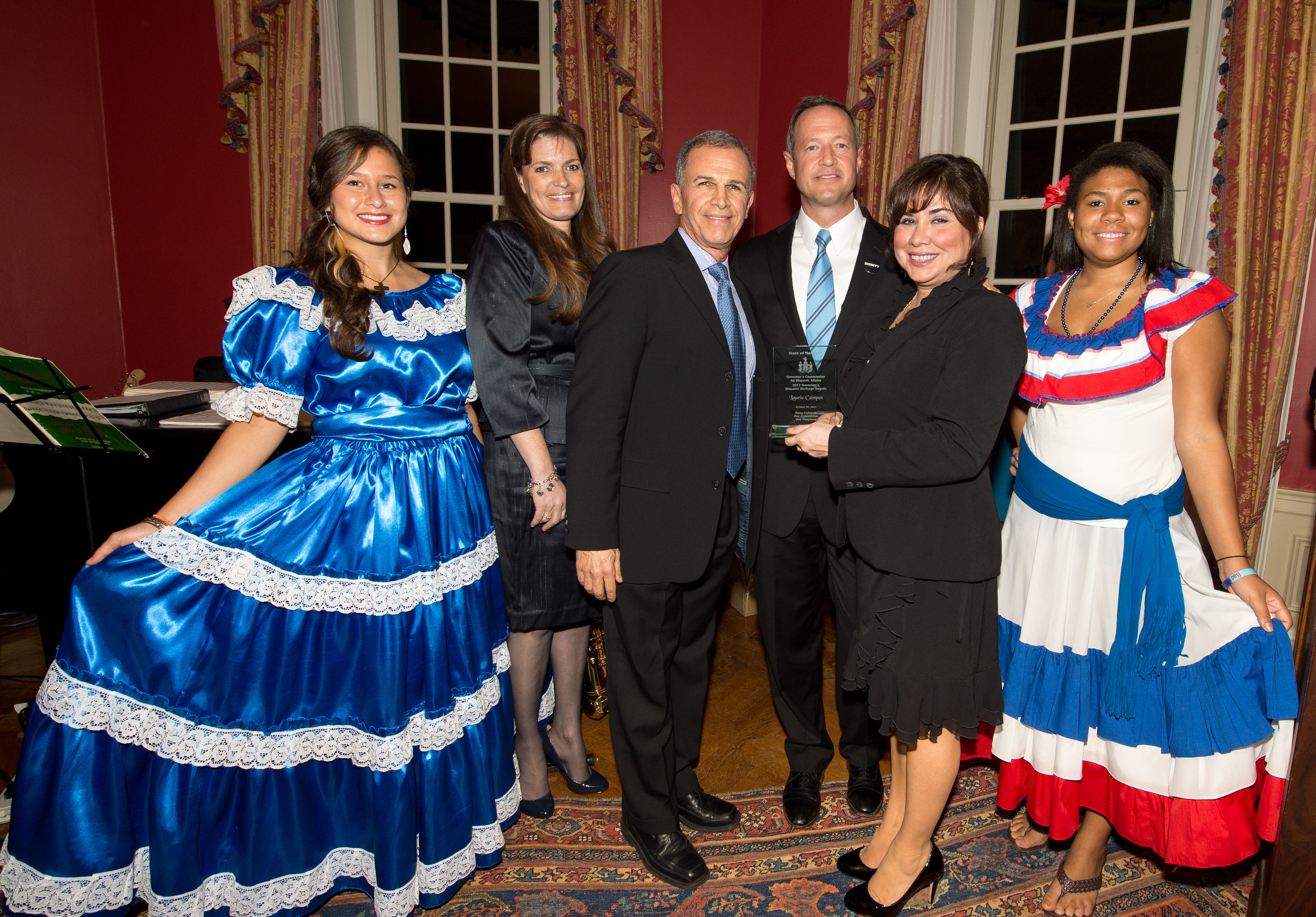 Six people pose at a Hispanic Heritage Month event, two wearing colorful traditional folk dresses.