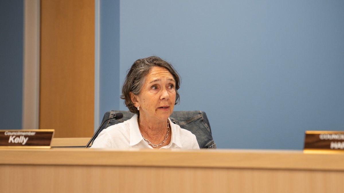 A woman seated at a government meeting dais with a nameplate reading Kelly, wearing a white blouse.