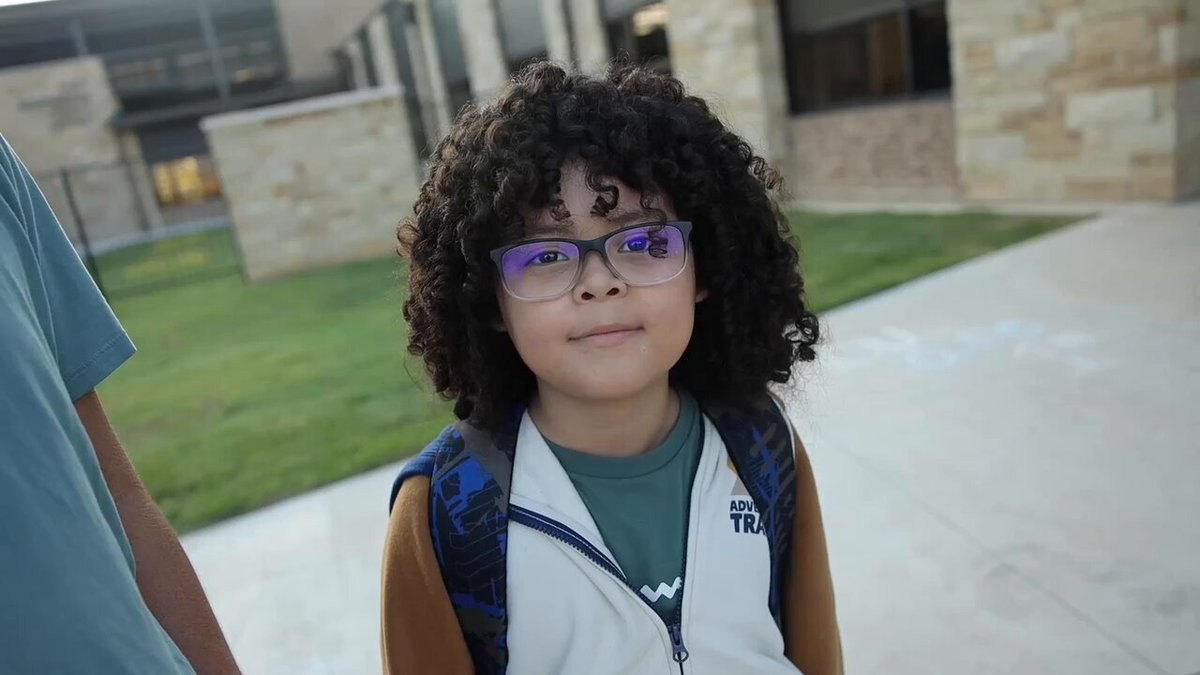 A smiling child with curly hair and purple glasses wears a backpack outdoors.