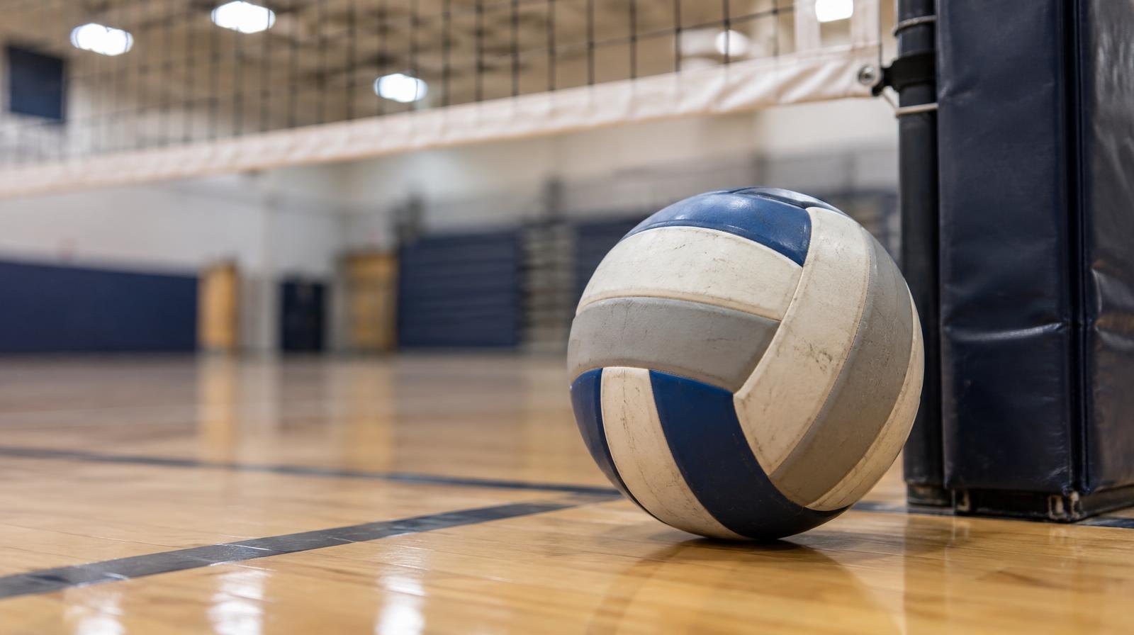 A volleyball resting on a hardwood gymnasium floor near a net post.