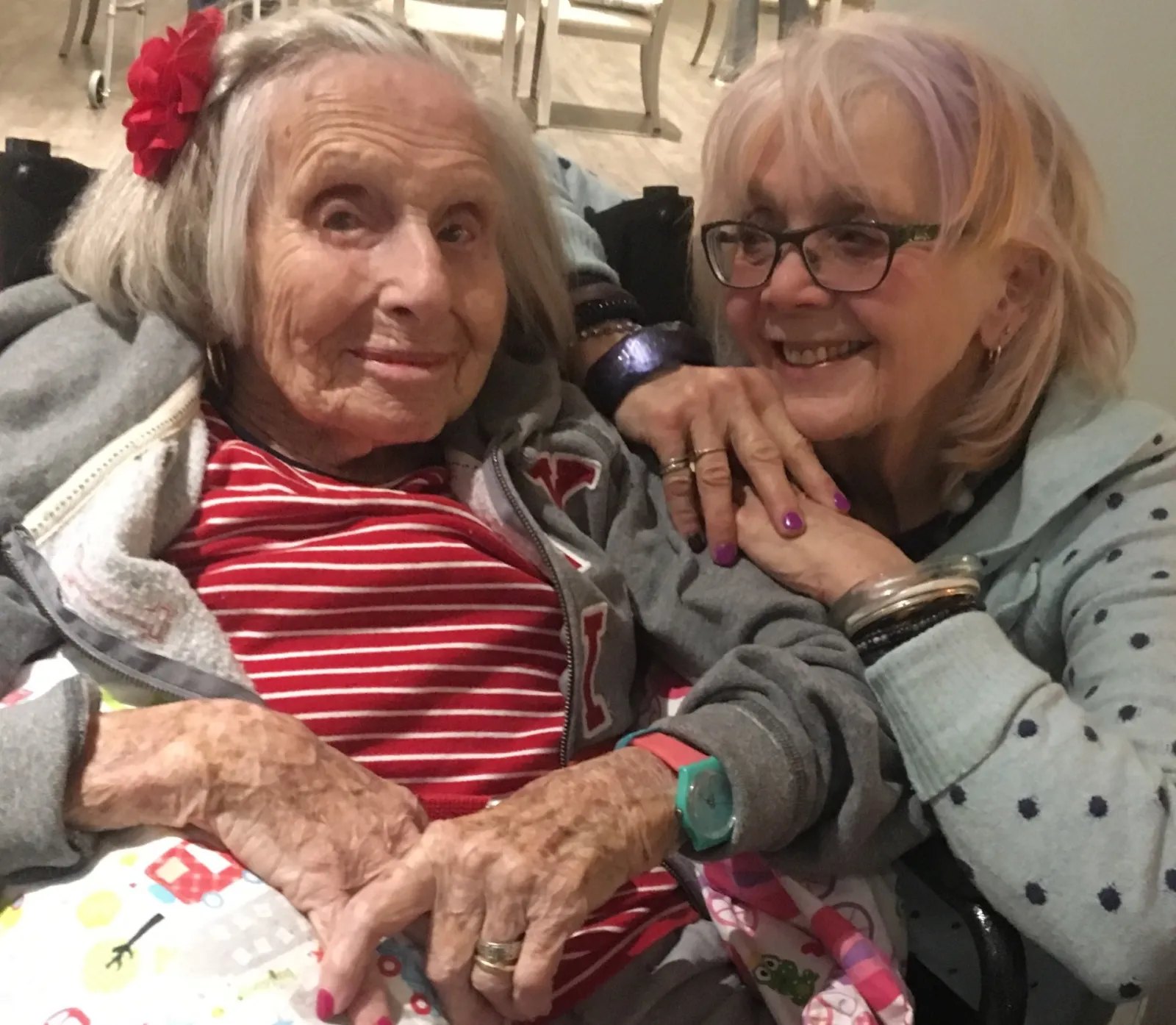 Two smiling women pose together, one elderly with white hair and a red flower accessory.