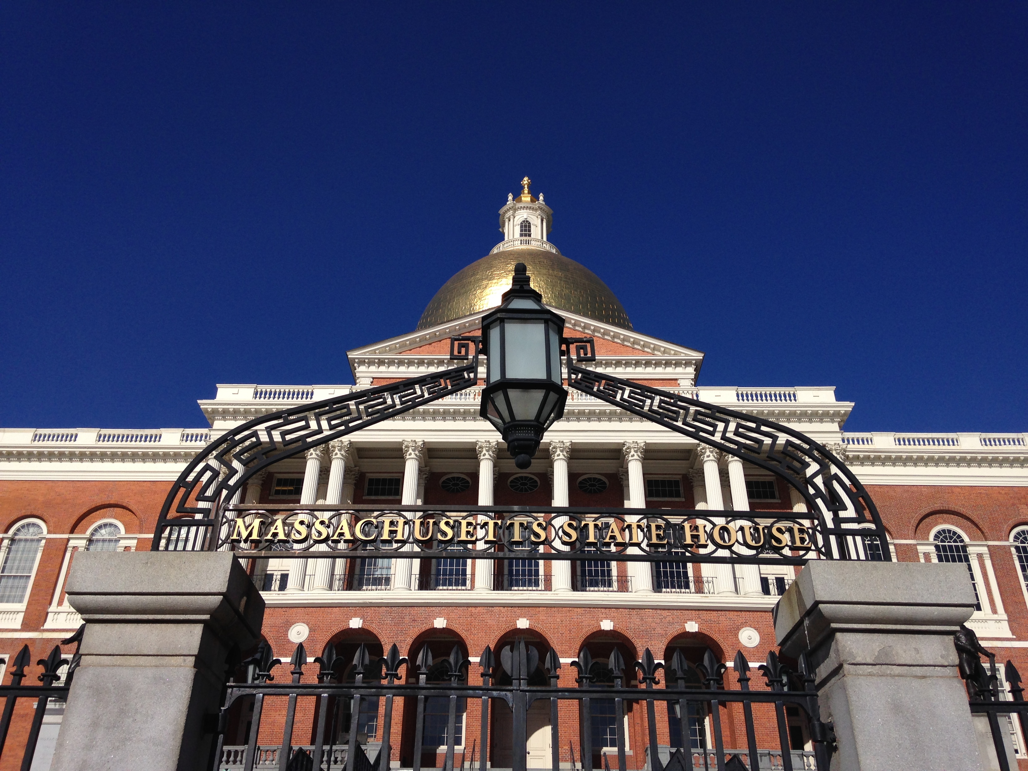 Gold-domed Massachusetts State House behind an ornate iron gate on a clear day.