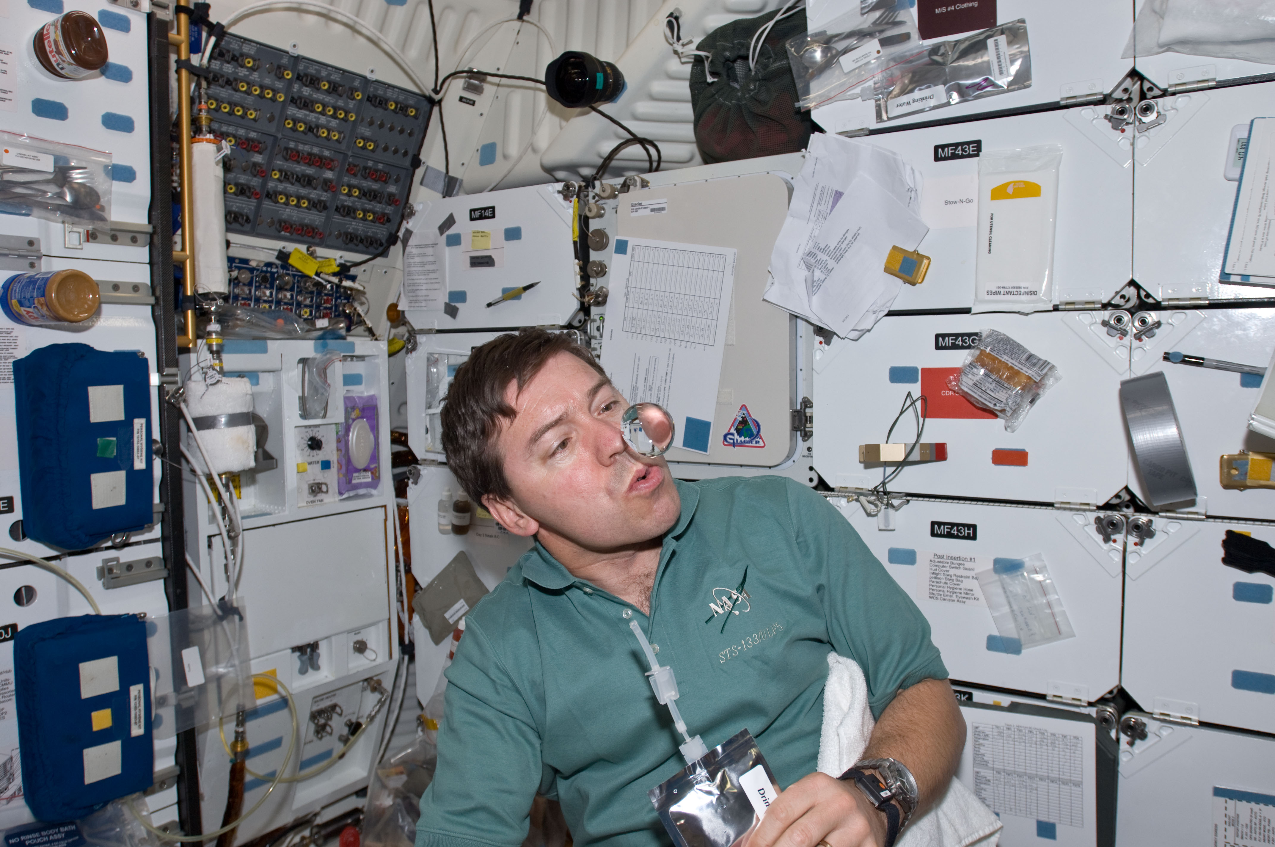 Astronaut in a green shirt watches a floating water bubble inside a spacecraft.