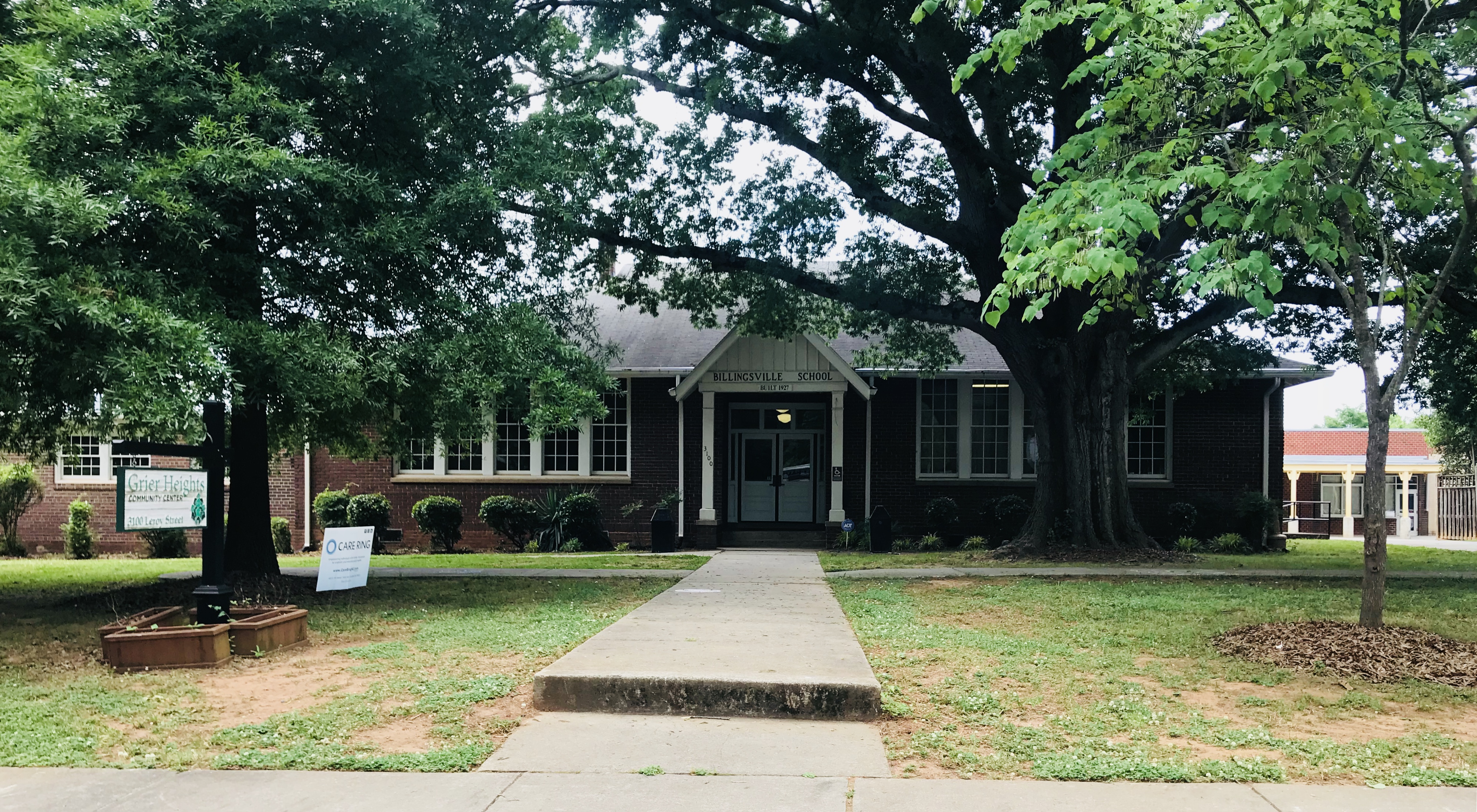 Single-story brick school building with mature trees and a concrete walkway to the entrance.