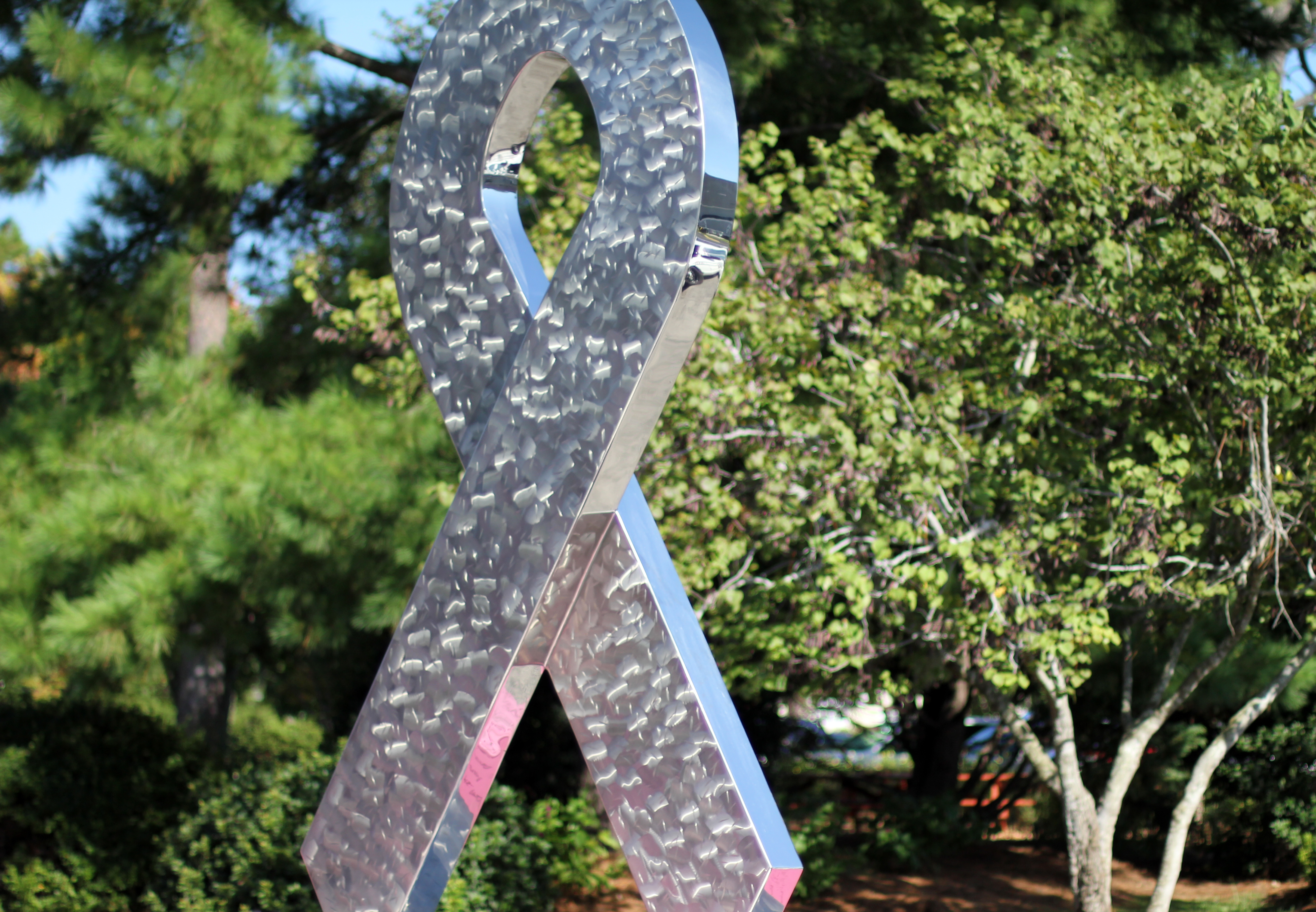 A large polished metal awareness ribbon sculpture standing outdoors among green trees.