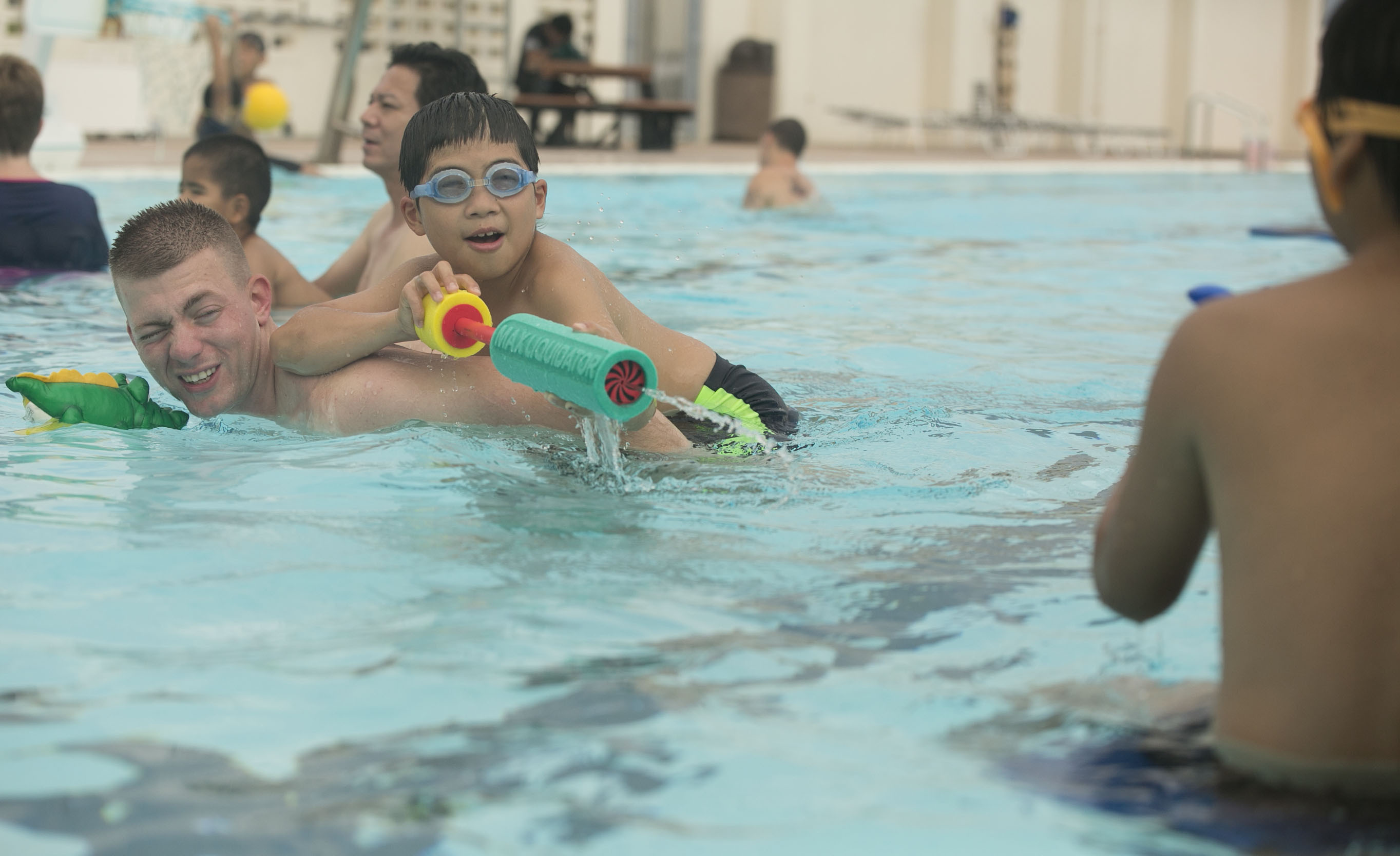 A boy in swim goggles rides on an adult's back in a pool while other children play nearby.