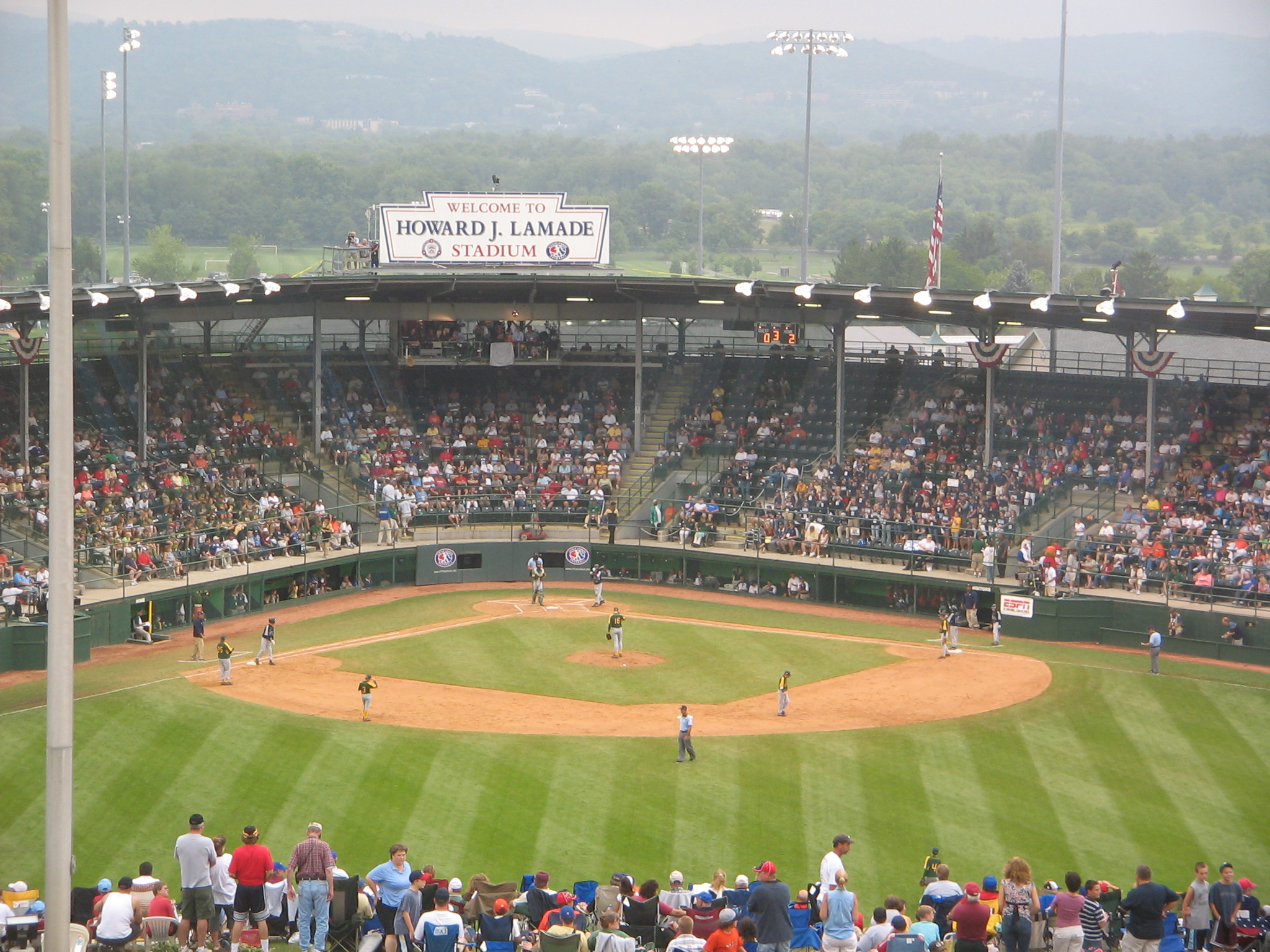 Little League World Series game in progress at a packed stadium with wooded hills beyond the outfield.