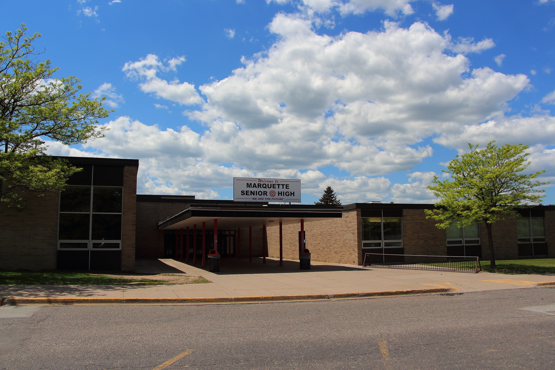 Front entrance of a low-rise brick high school building with a covered canopy and trees.