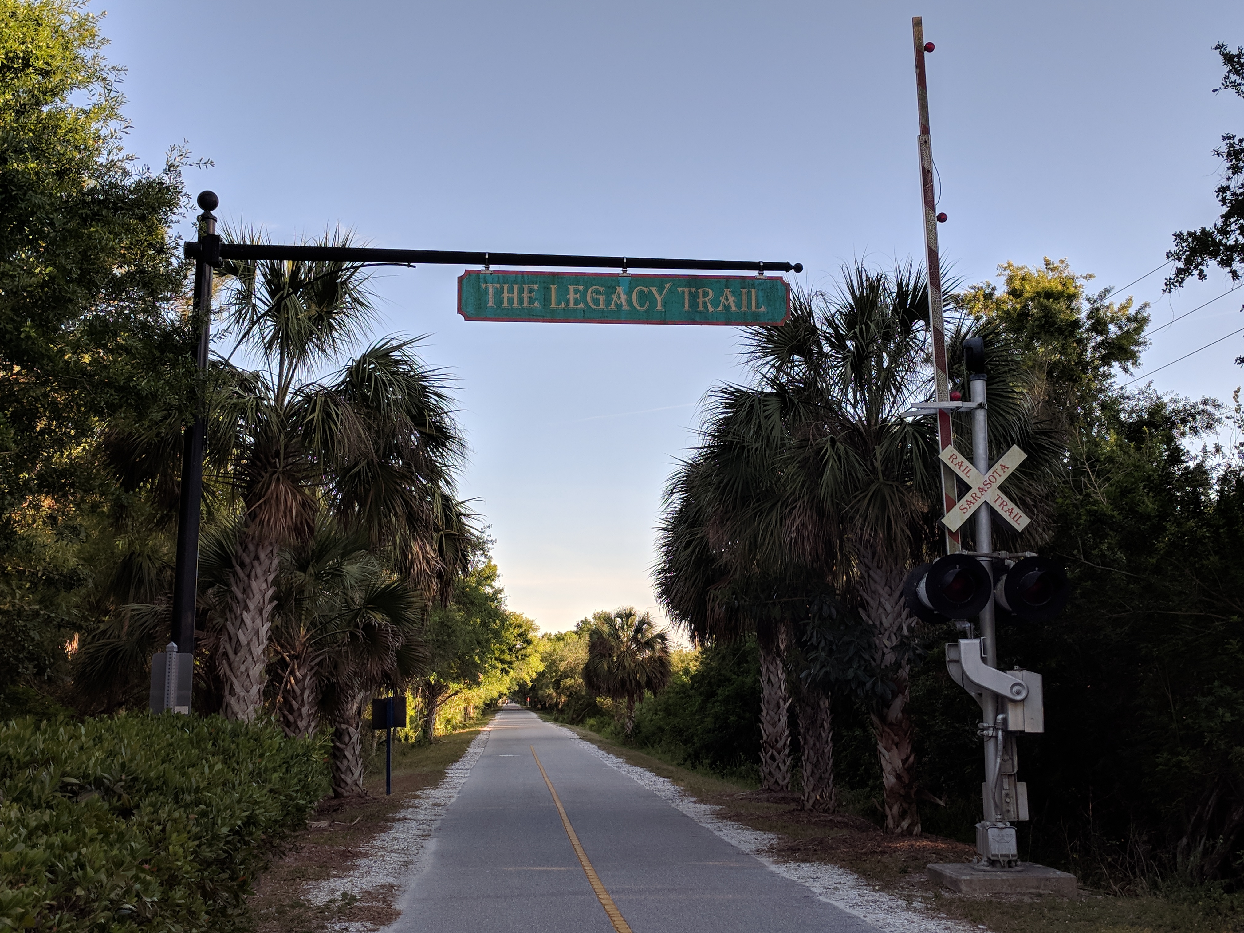 Green overhead Legacy Trail sign on a paved path flanked by palm trees.
