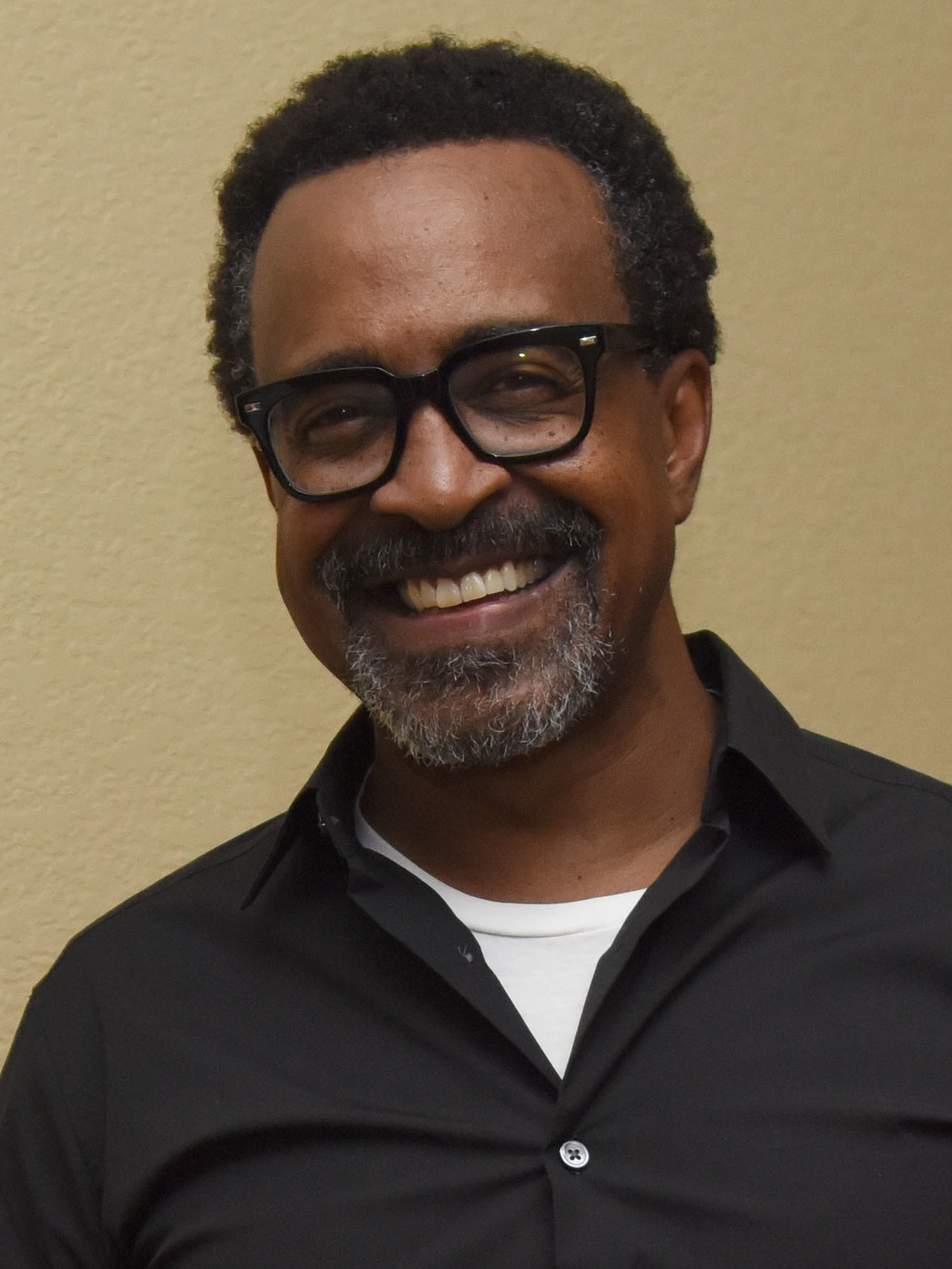 A smiling man wearing black-framed glasses and a black button-up shirt in a headshot-style portrait.