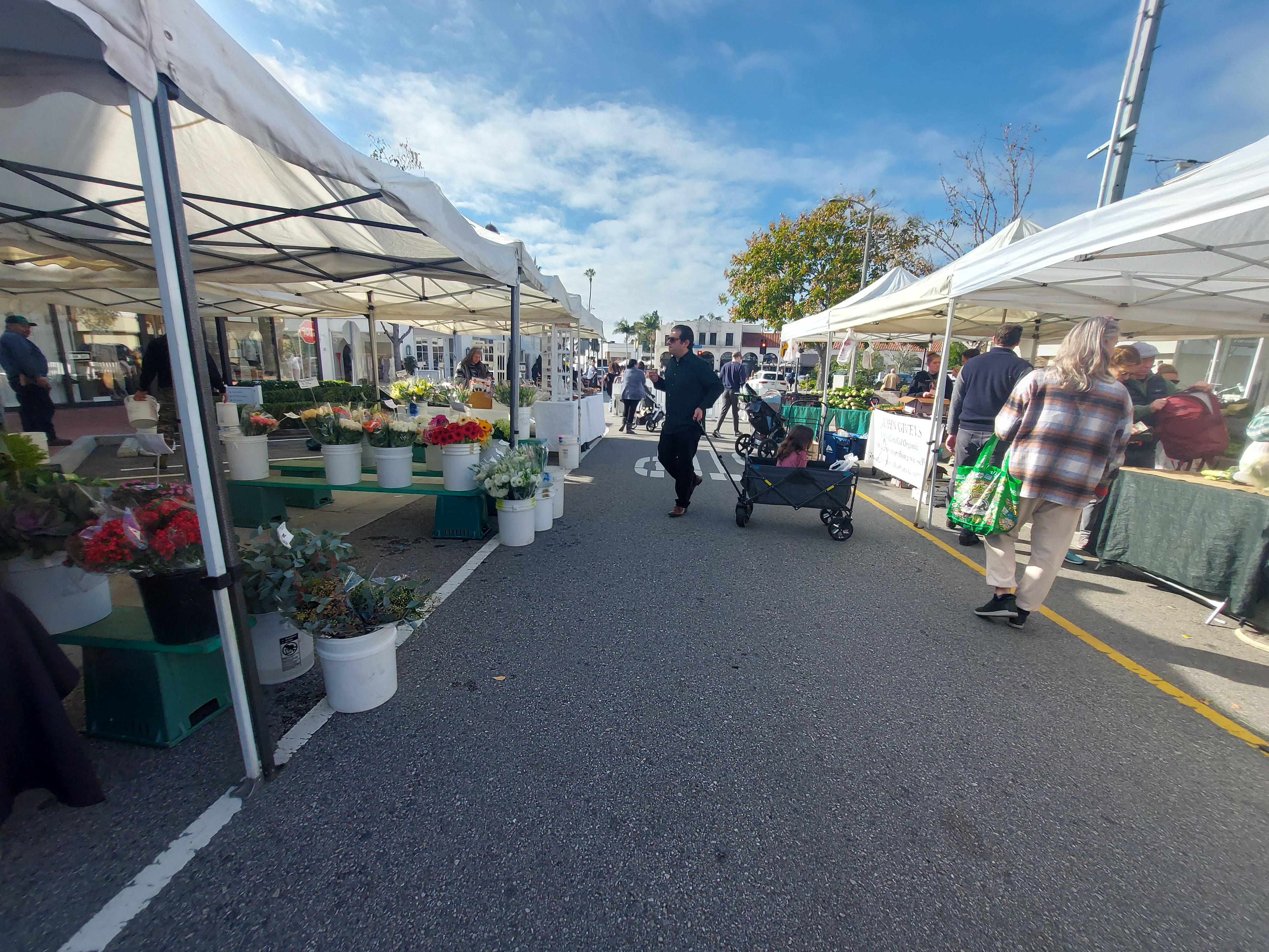 White canopy tents line a paved market aisle as shoppers browse vendor stalls.