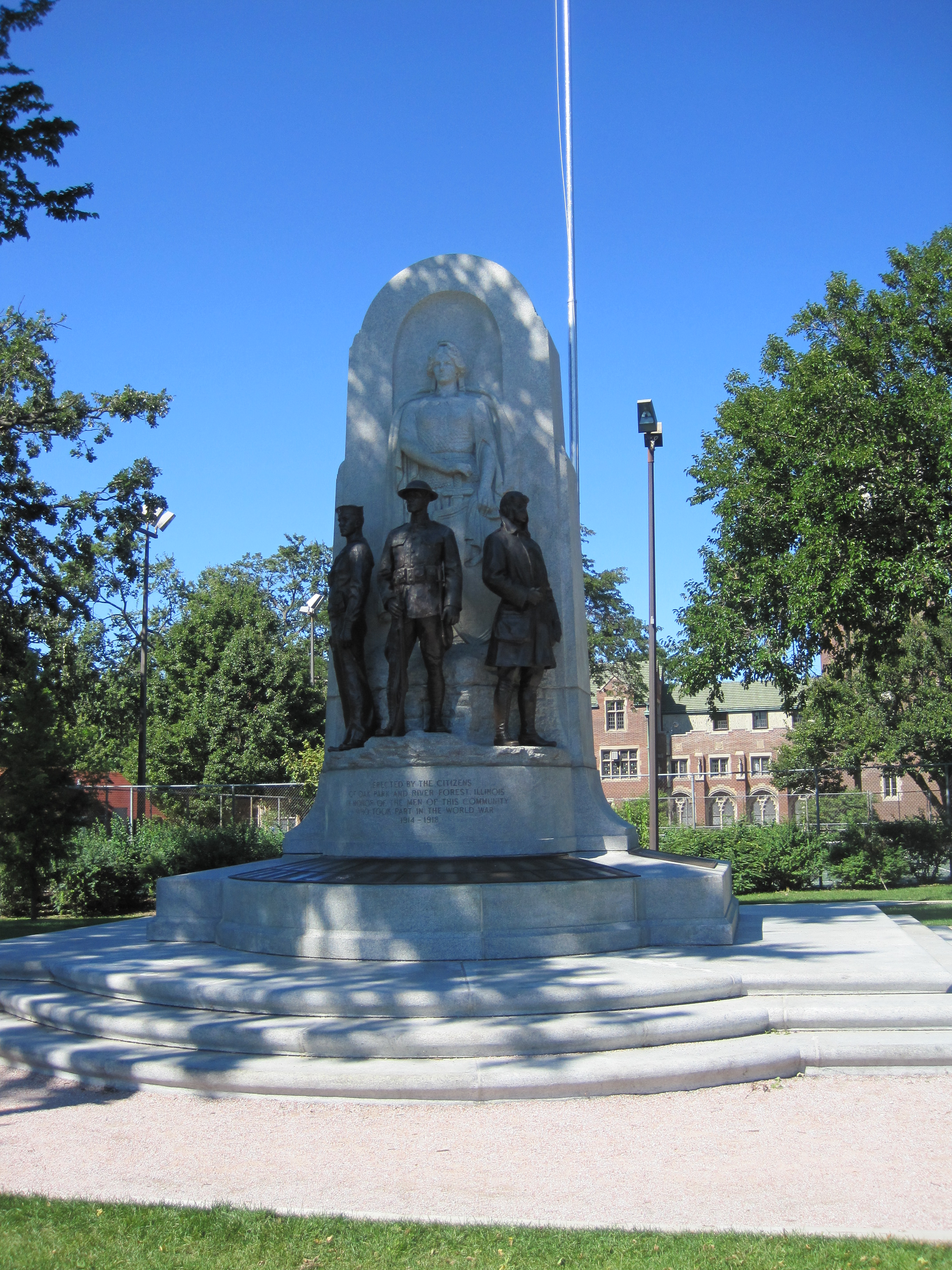 Stone arch war monument with bronze soldier figures in a park, trees and brick building behind.