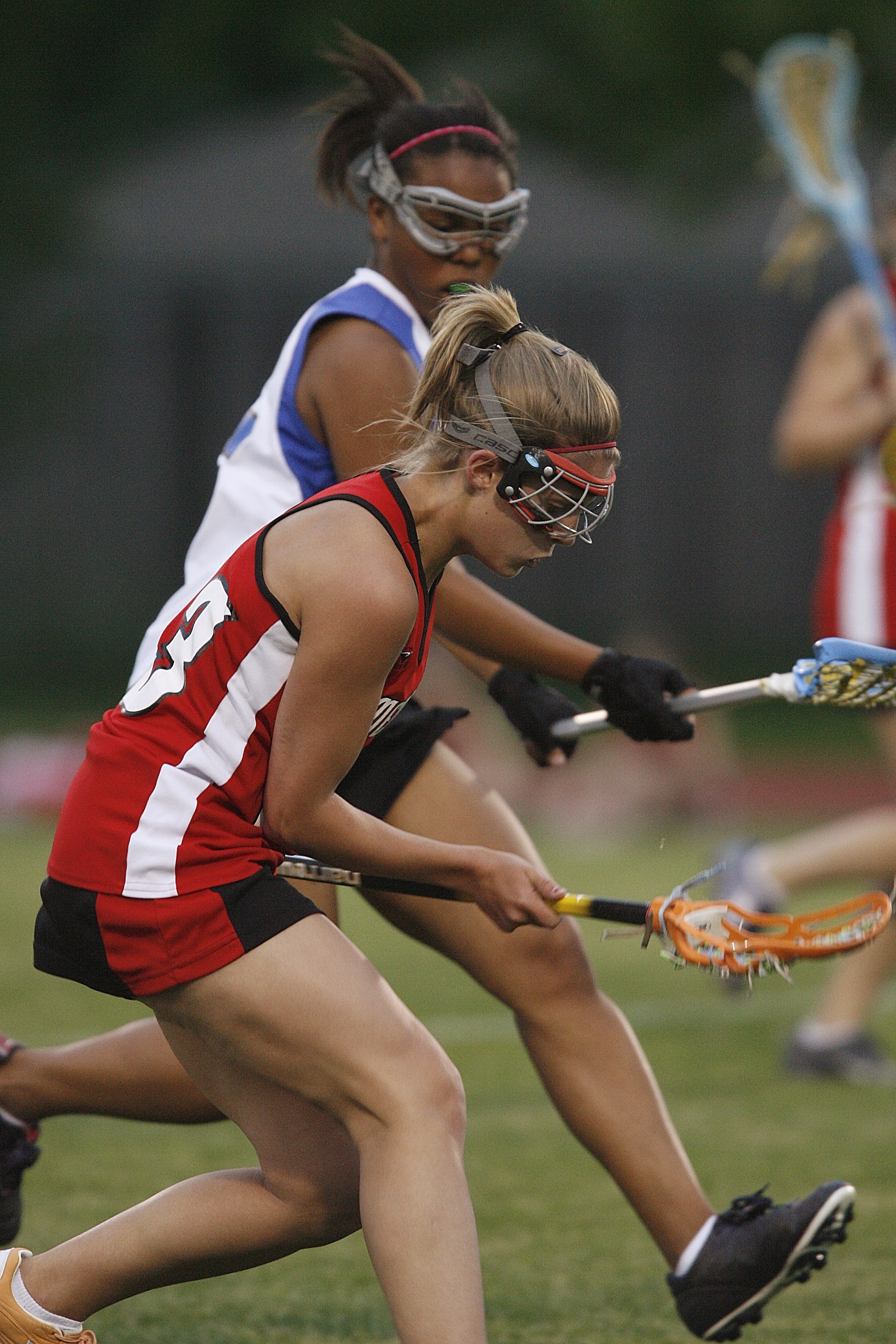 Two female lacrosse players run on a field during a game, both carrying lacrosse sticks.