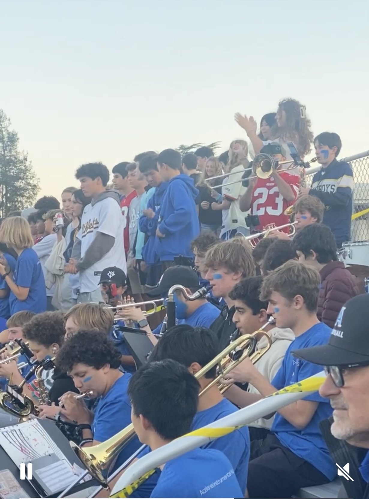 Students in blue uniforms playing trombones and saxophones in stadium bleachers at dusk.