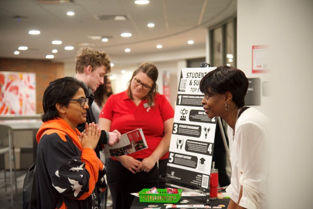 Three people in conversation at an information table inside a college building.