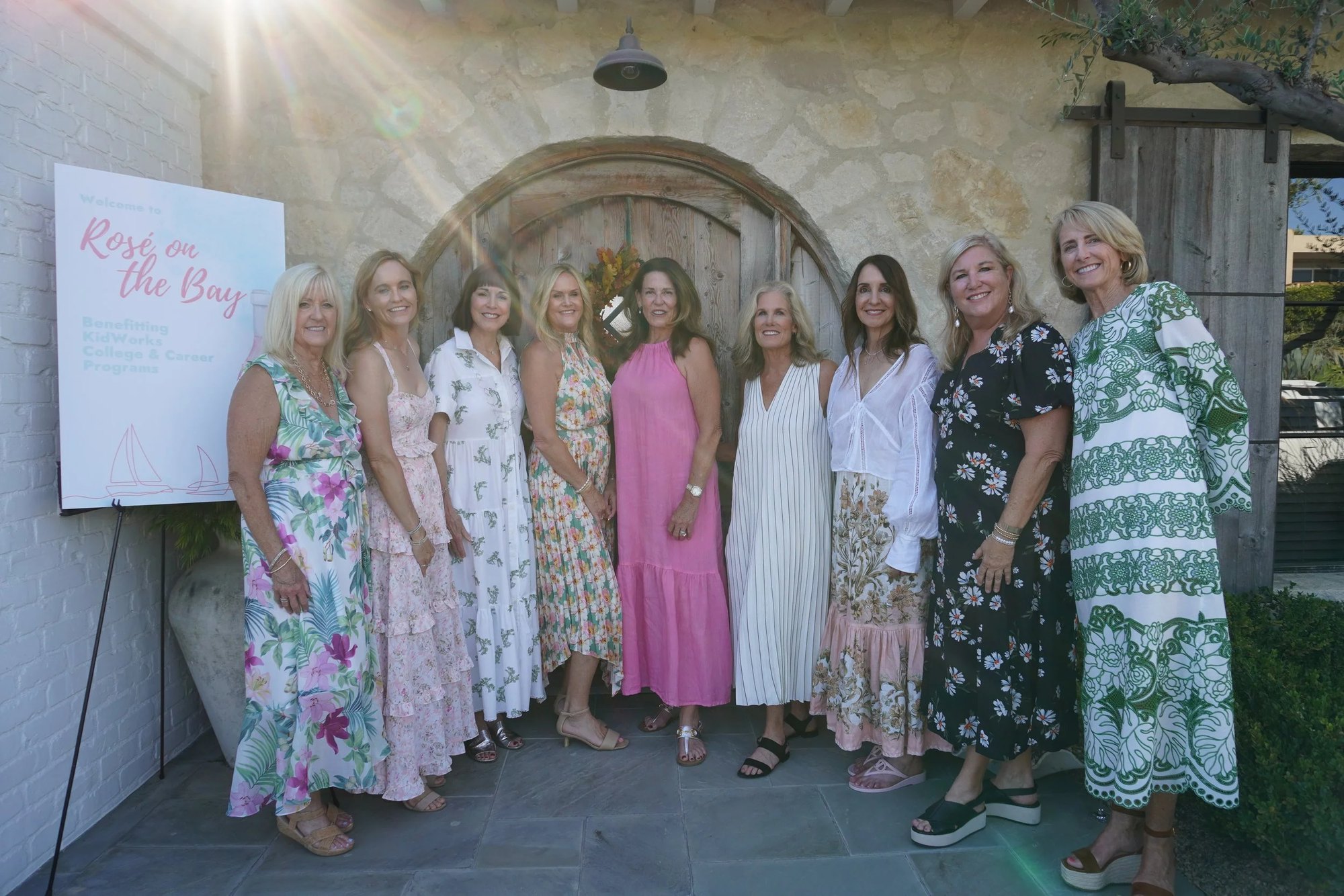 Nine women in summer dresses posing together beside a Rosé on the Bay sign in front of a stone archway.