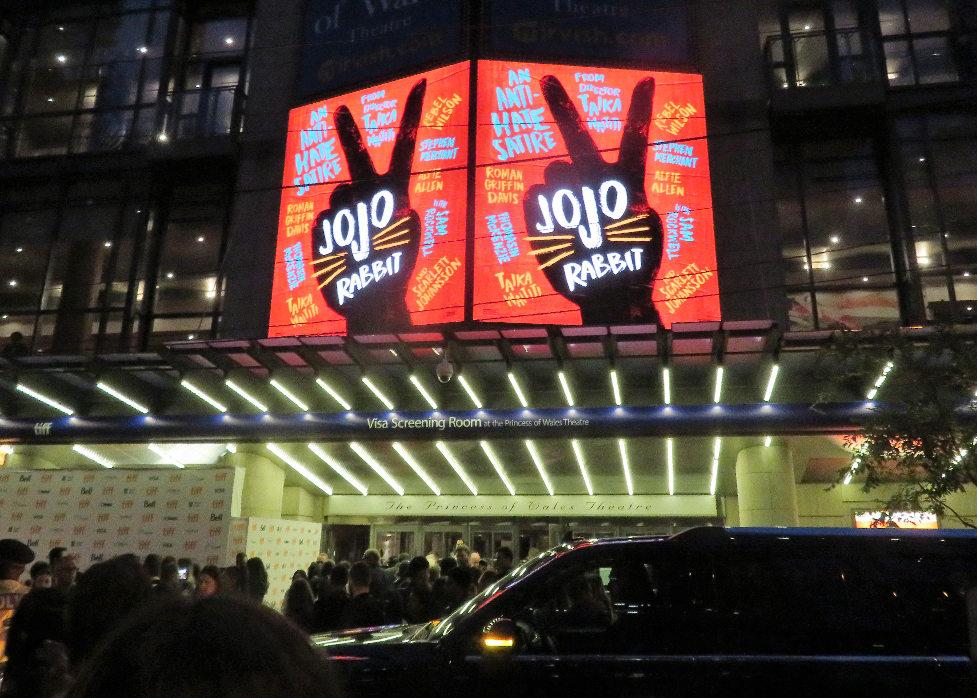 Theater marquee lit up with Jojo Rabbit signage at night, crowd gathered outside.