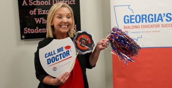 Smiling woman holds a 'Call Me Doctor' sign in front of a Georgia's BEST banner.