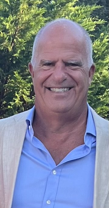Smiling man with gray hair in a blazer standing outdoors among green foliage.