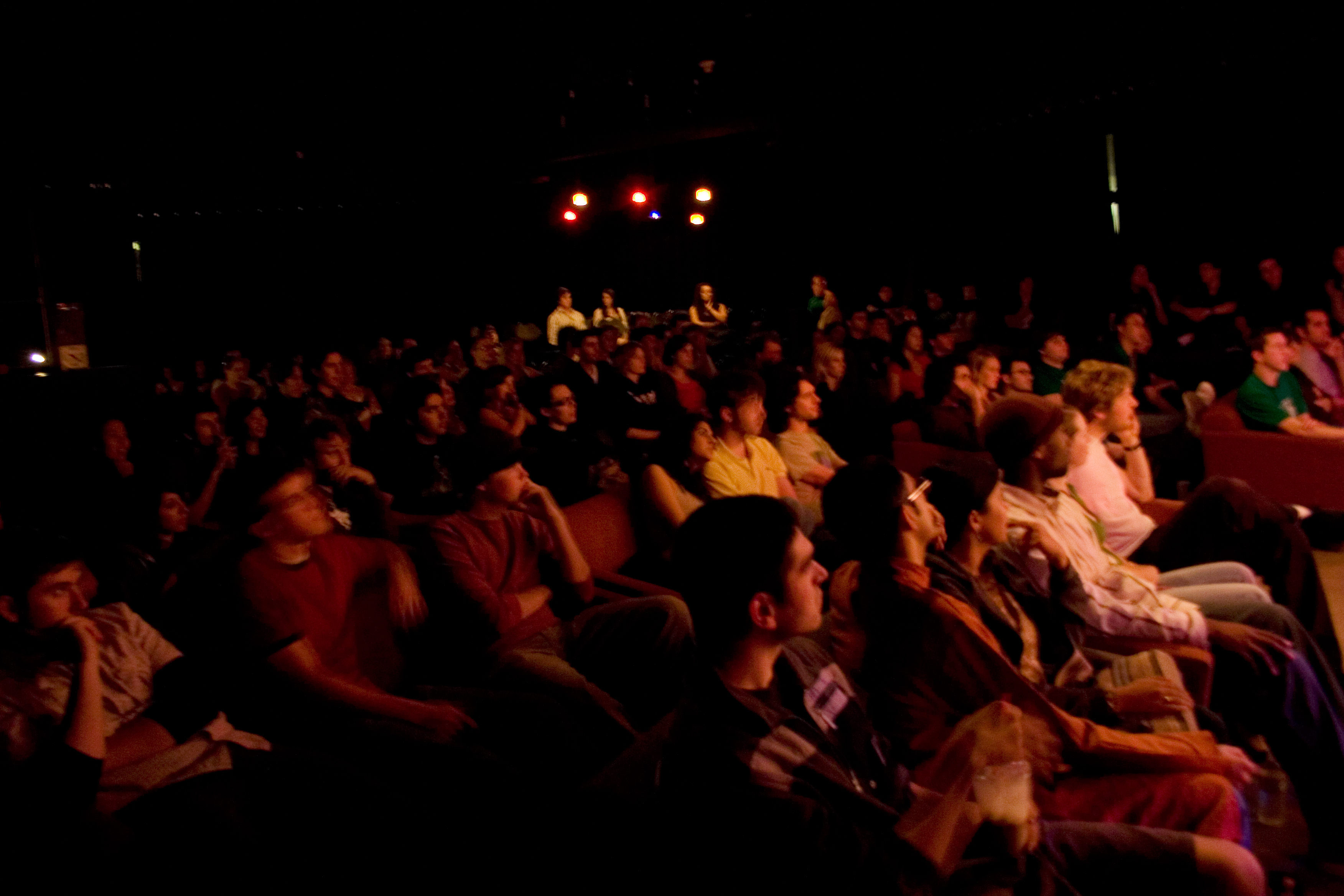 Audience seated in rows in a dimly lit performance space with stage lights in the background.