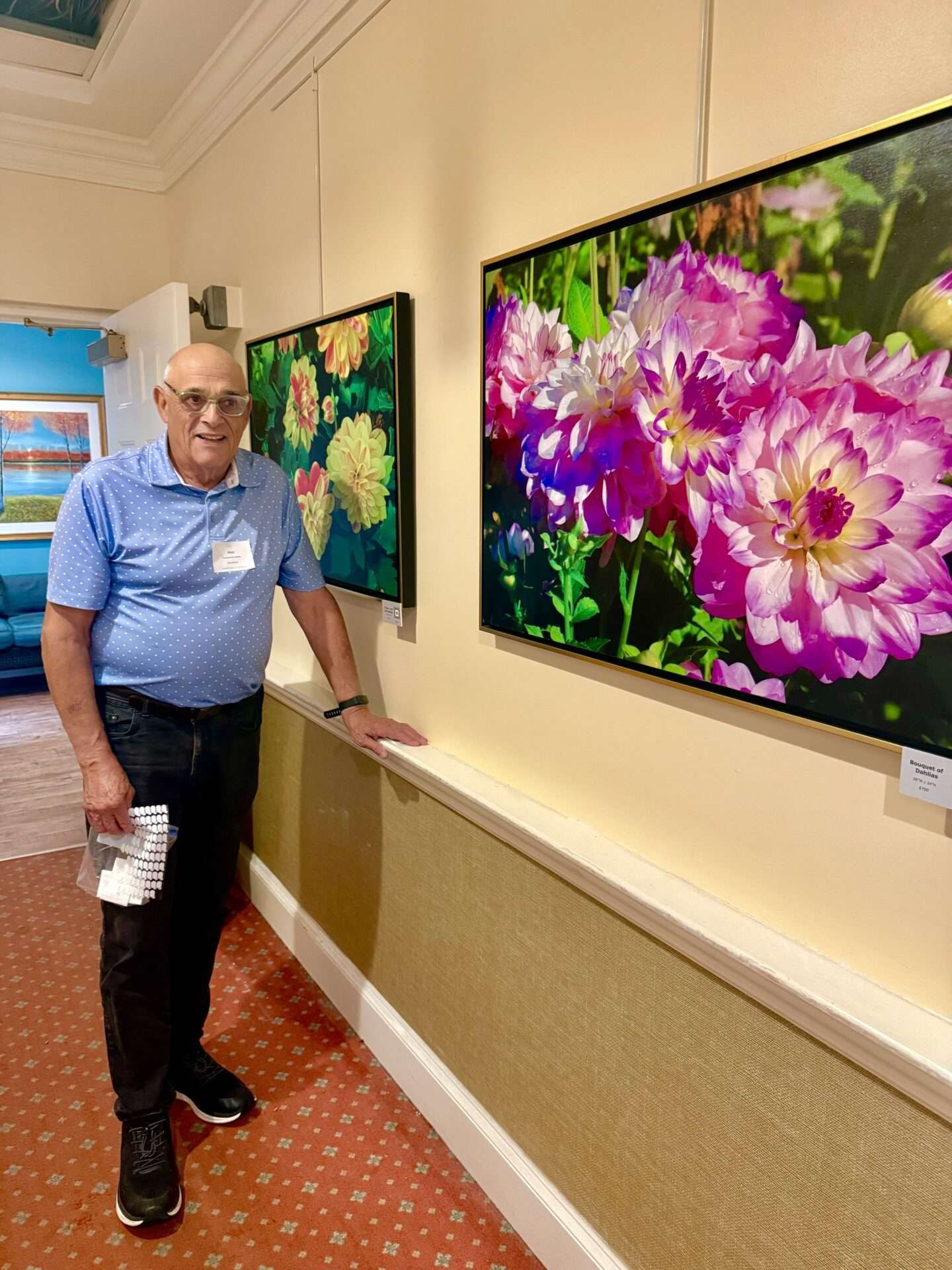 A man in a blue polo shirt stands next to two large framed floral photographs on a beige wall.