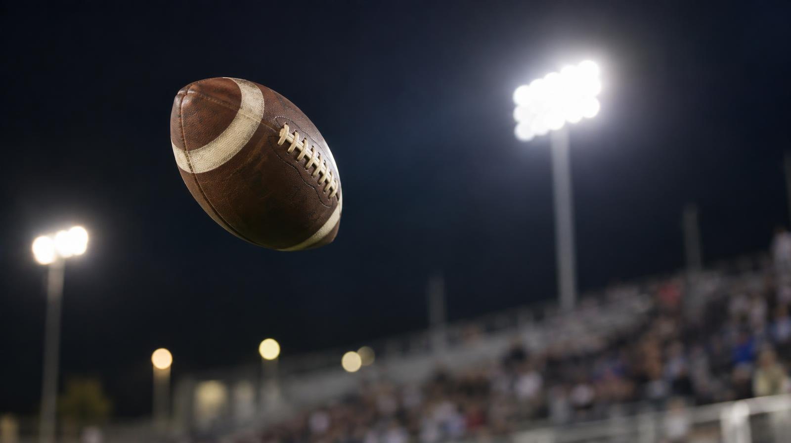 An American football spiraling through the air with bright stadium lights in the background.