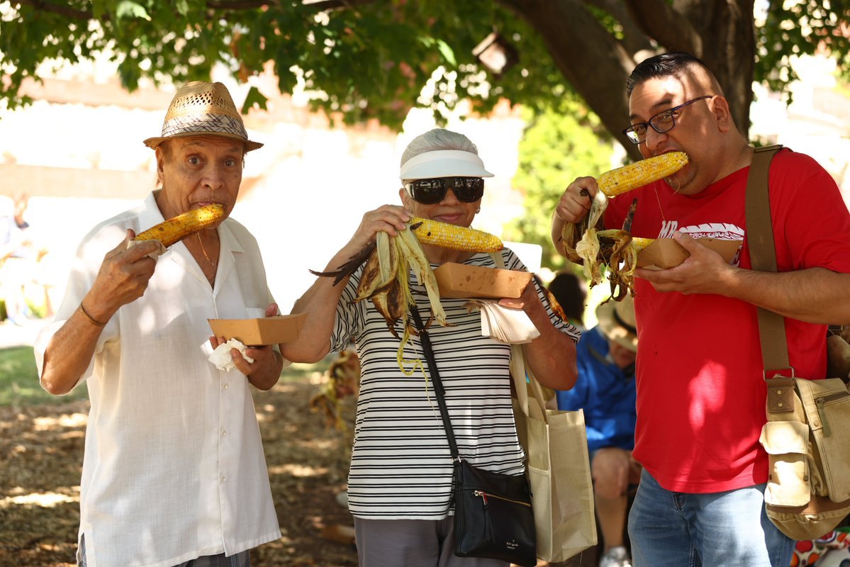 Three people stand outdoors eating roasted corn on the cob.