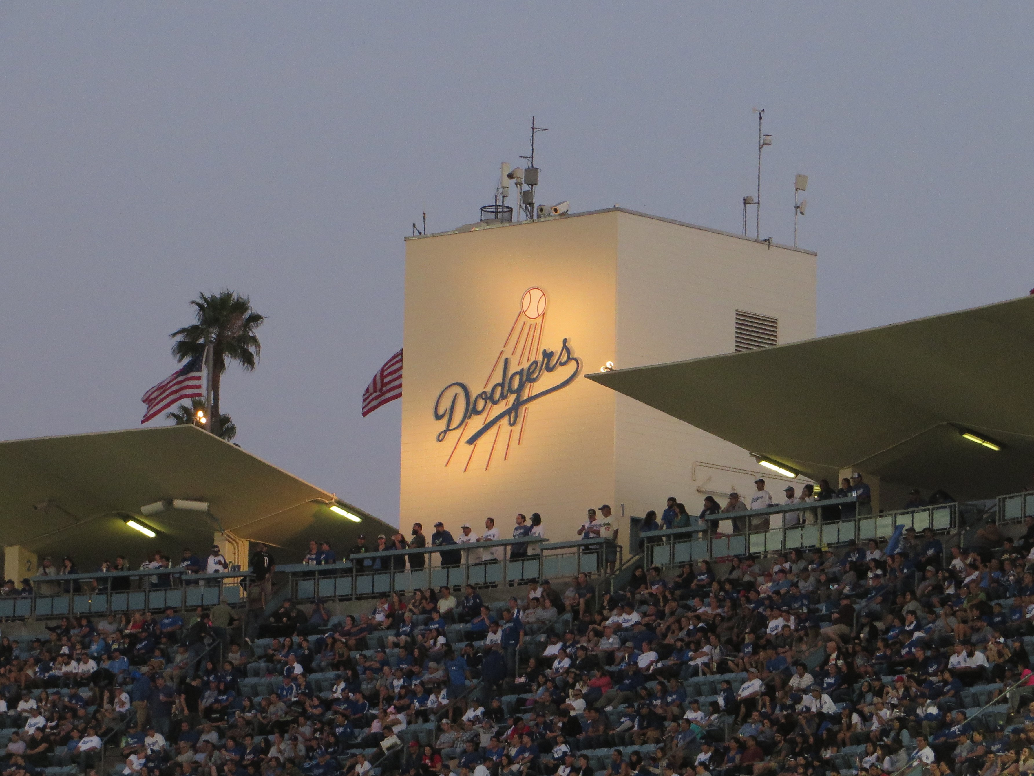 Illuminated Dodgers logo tower at Dodger Stadium at dusk with fans in the stands.