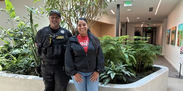 A sheriff's deputy and a woman in casual clothes smile together indoors.
