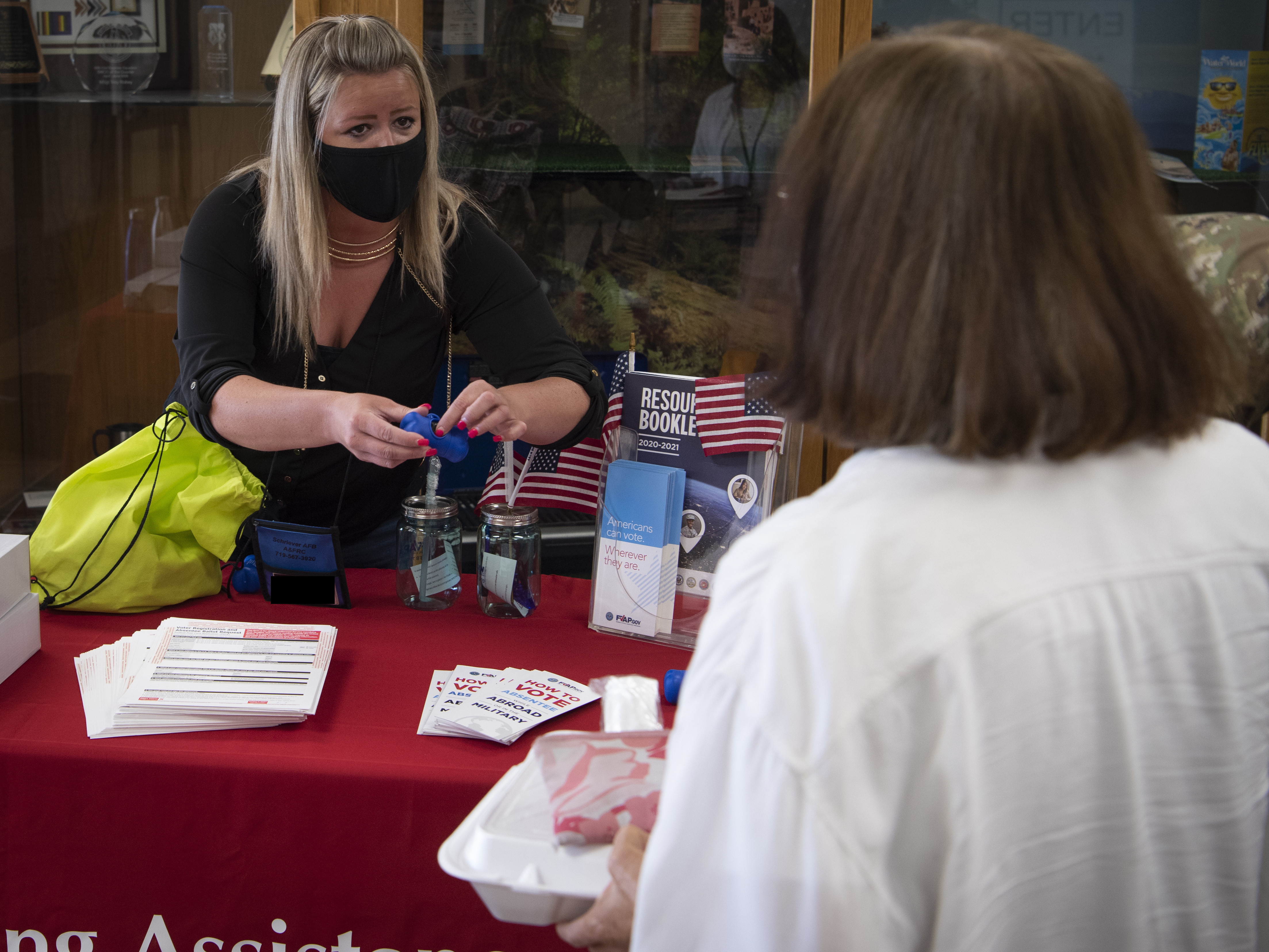A masked woman at a red-draped table hands materials to a visitor at a voting assistance booth.