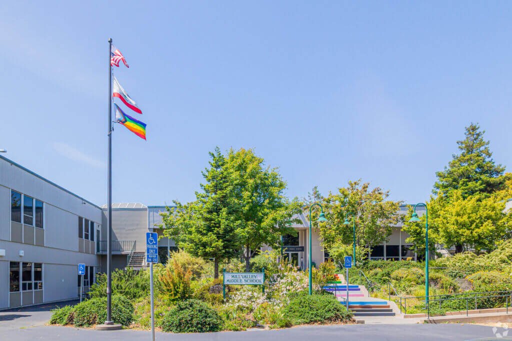 Exterior of a school campus with a flagpole flying three flags and low-rise buildings.