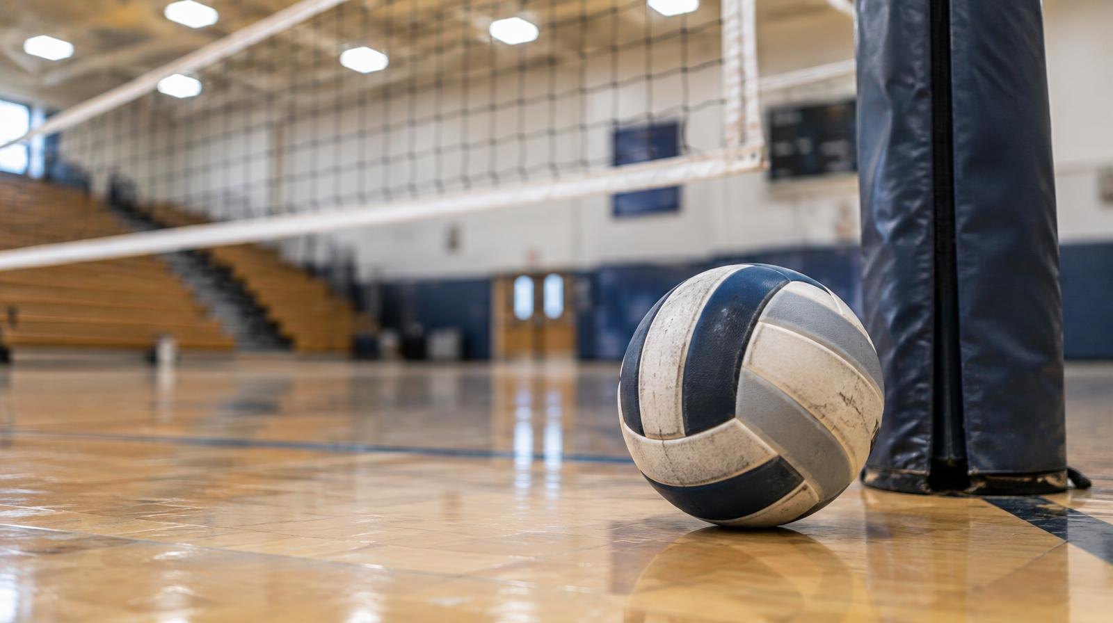 A volleyball resting on a hardwood floor near a volleyball net post.