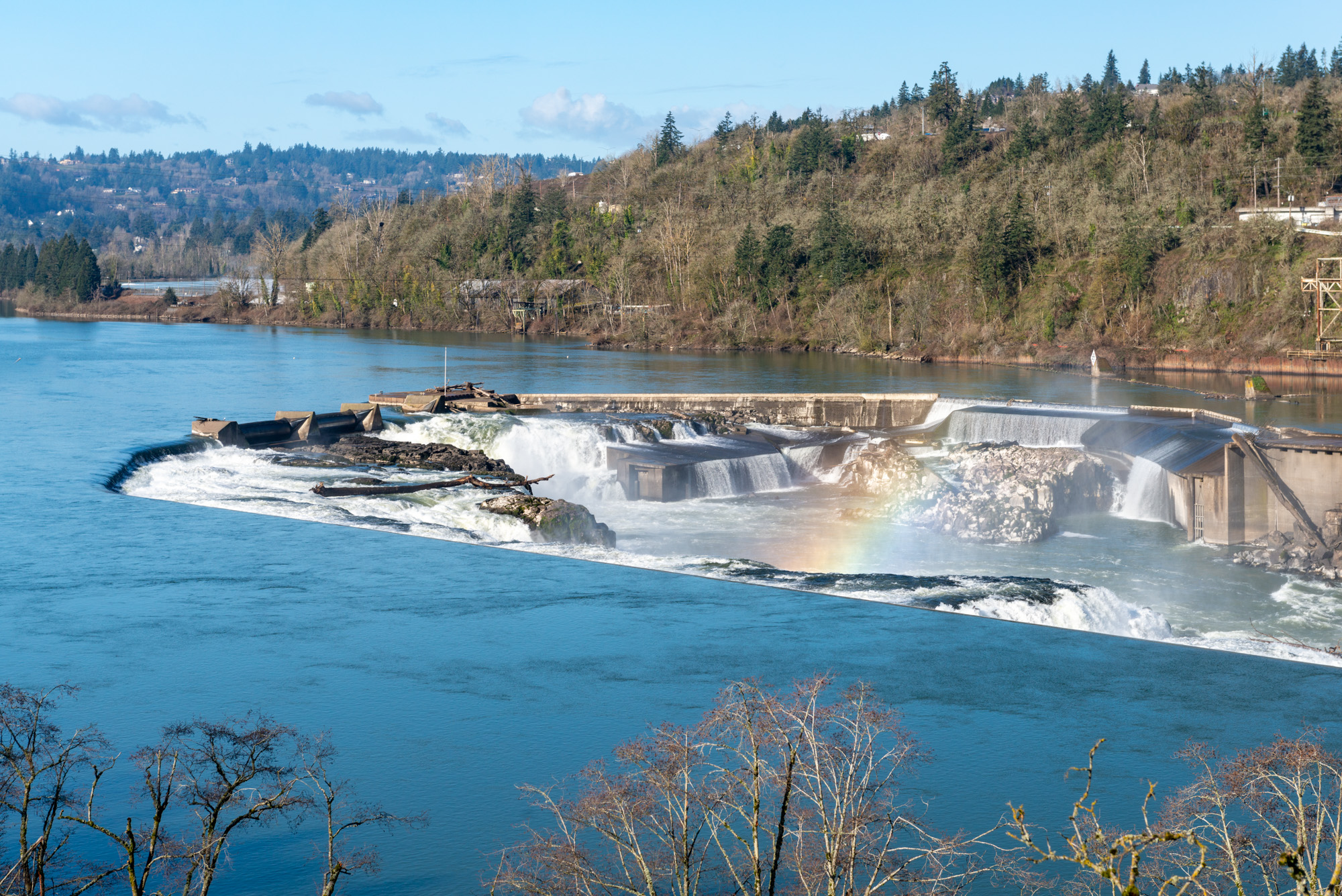 Wide view of Willamette Falls with a rainbow visible in the mist and forested hillsides behind.