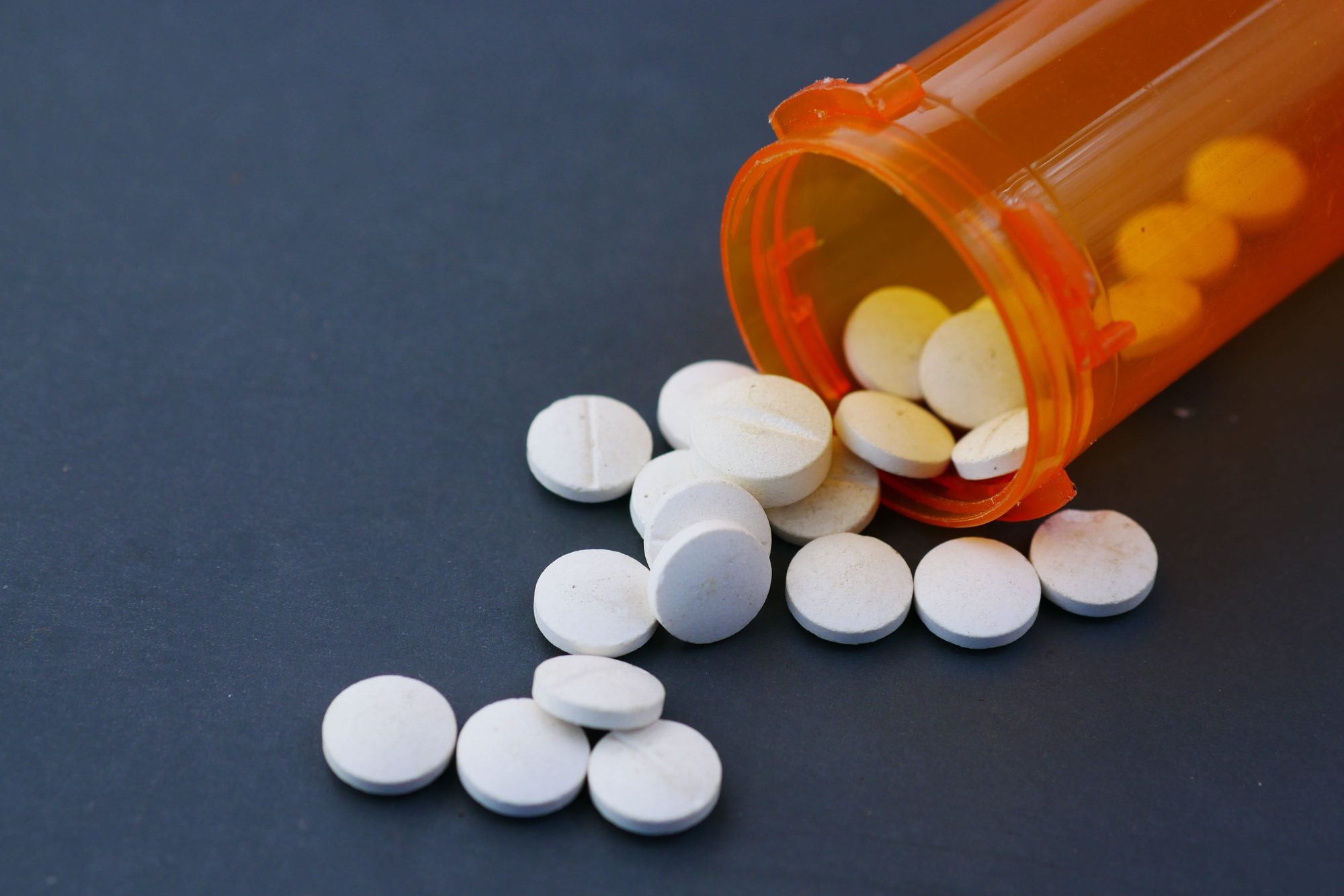 Close-up of white pills scattered from an orange prescription bottle on a gray surface.