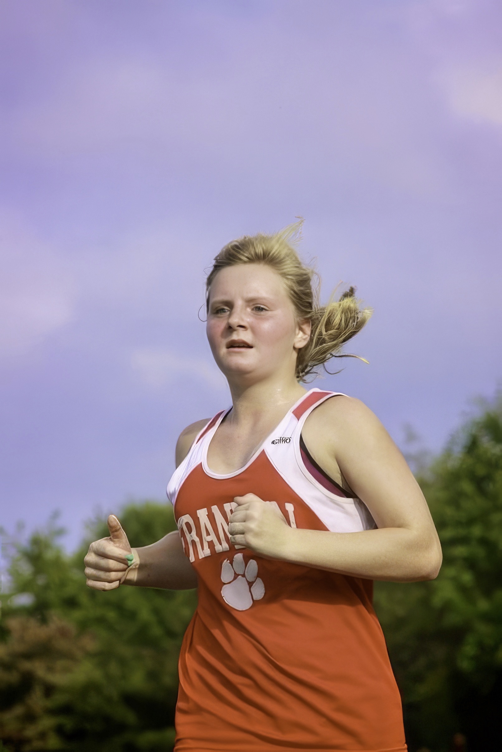 A female high school runner in an orange sleeveless jersey races outdoors with trees behind her.