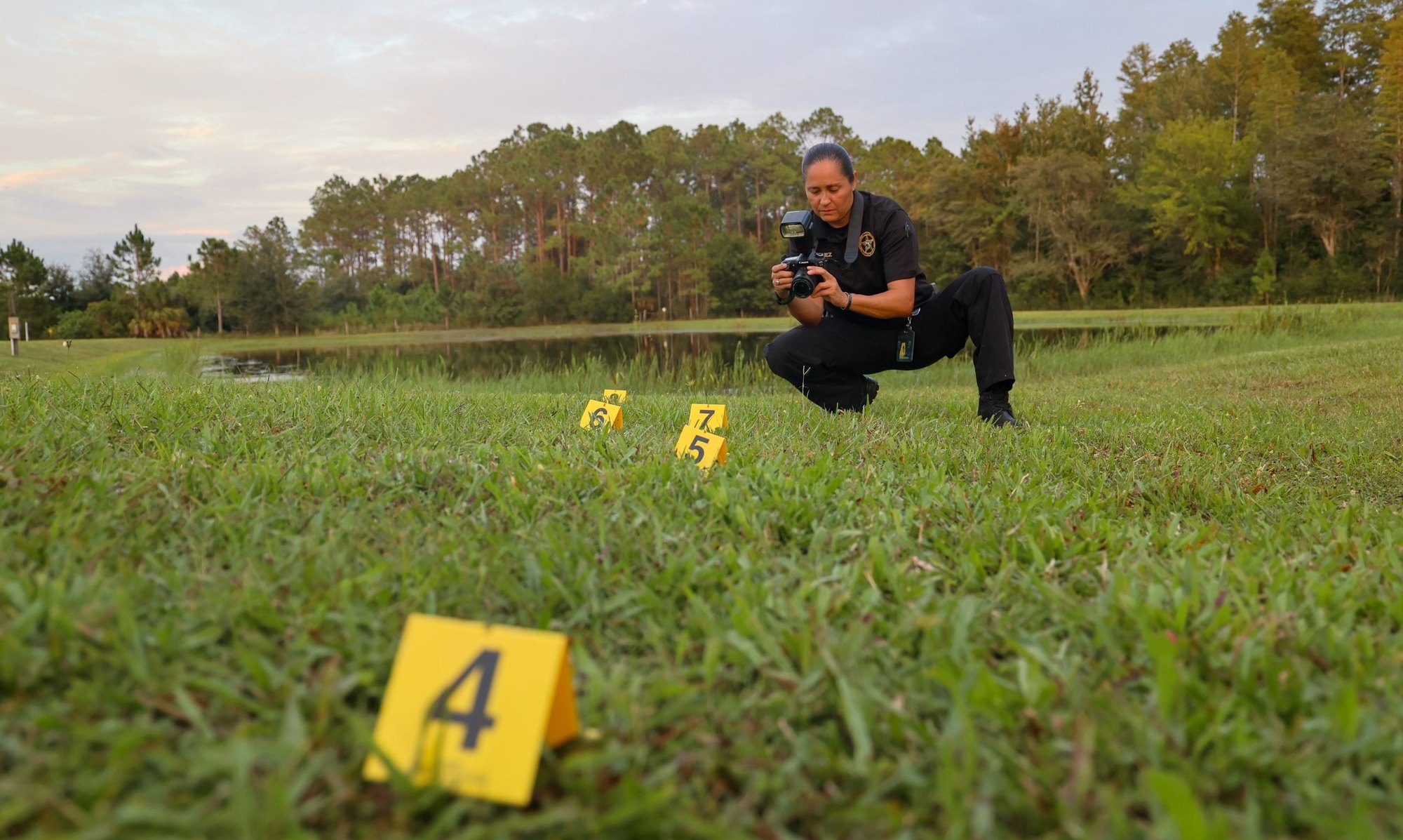 Law enforcement officer crouching to photograph yellow numbered evidence markers on grass.