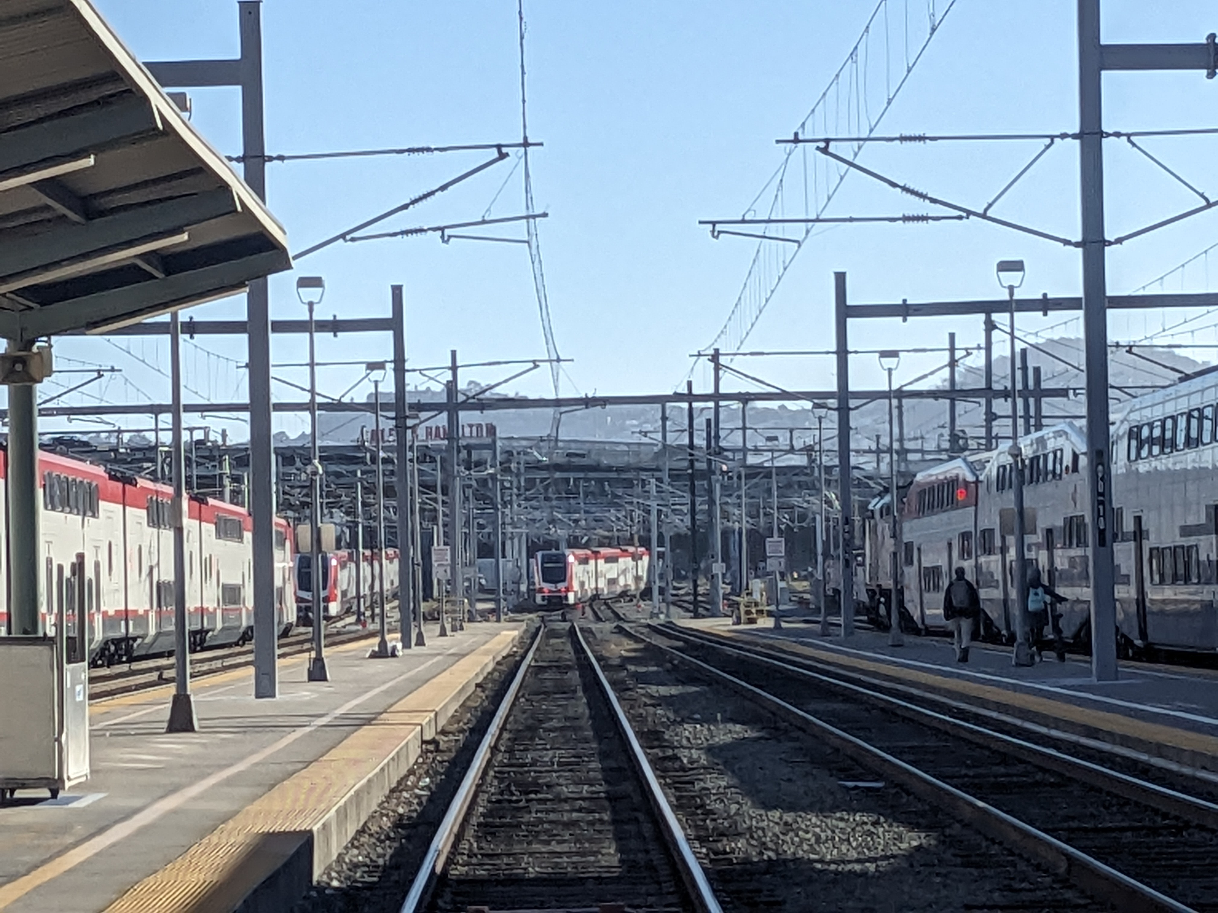 A Caltrain electric train at a station with overhead wires and covered platforms visible.