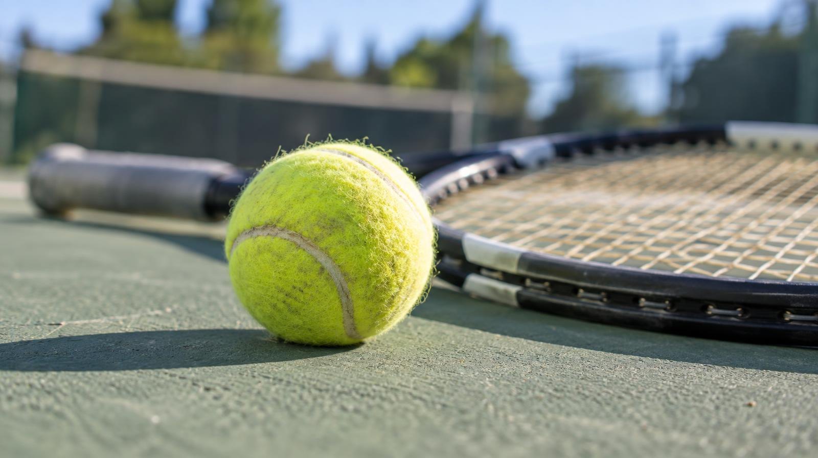 Yellow tennis ball resting beside a racket on a green hard court.