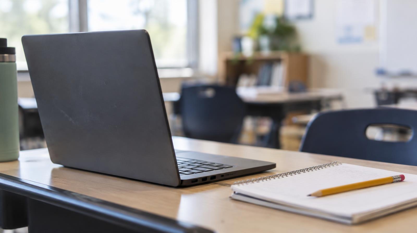 Open laptop computer on a wooden school desk beside a pencil and notebook.