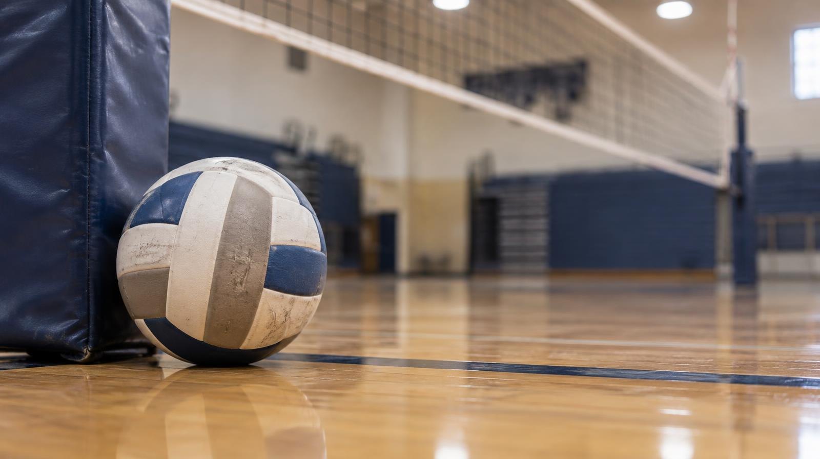 A volleyball resting on a hardwood gymnasium floor near a net post.