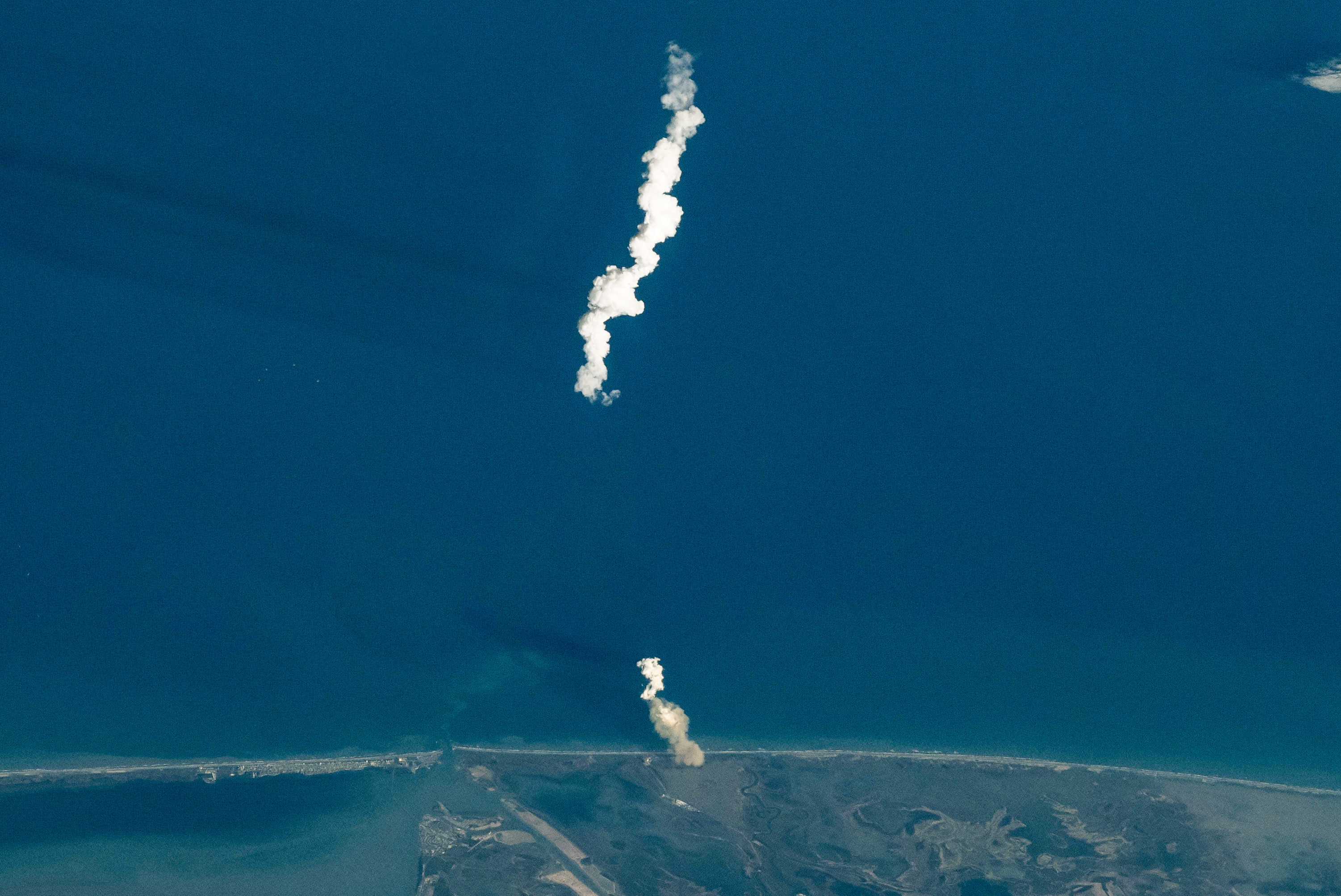 Rocket launch plume rising from a coastal launch site, seen from orbit above Earth.