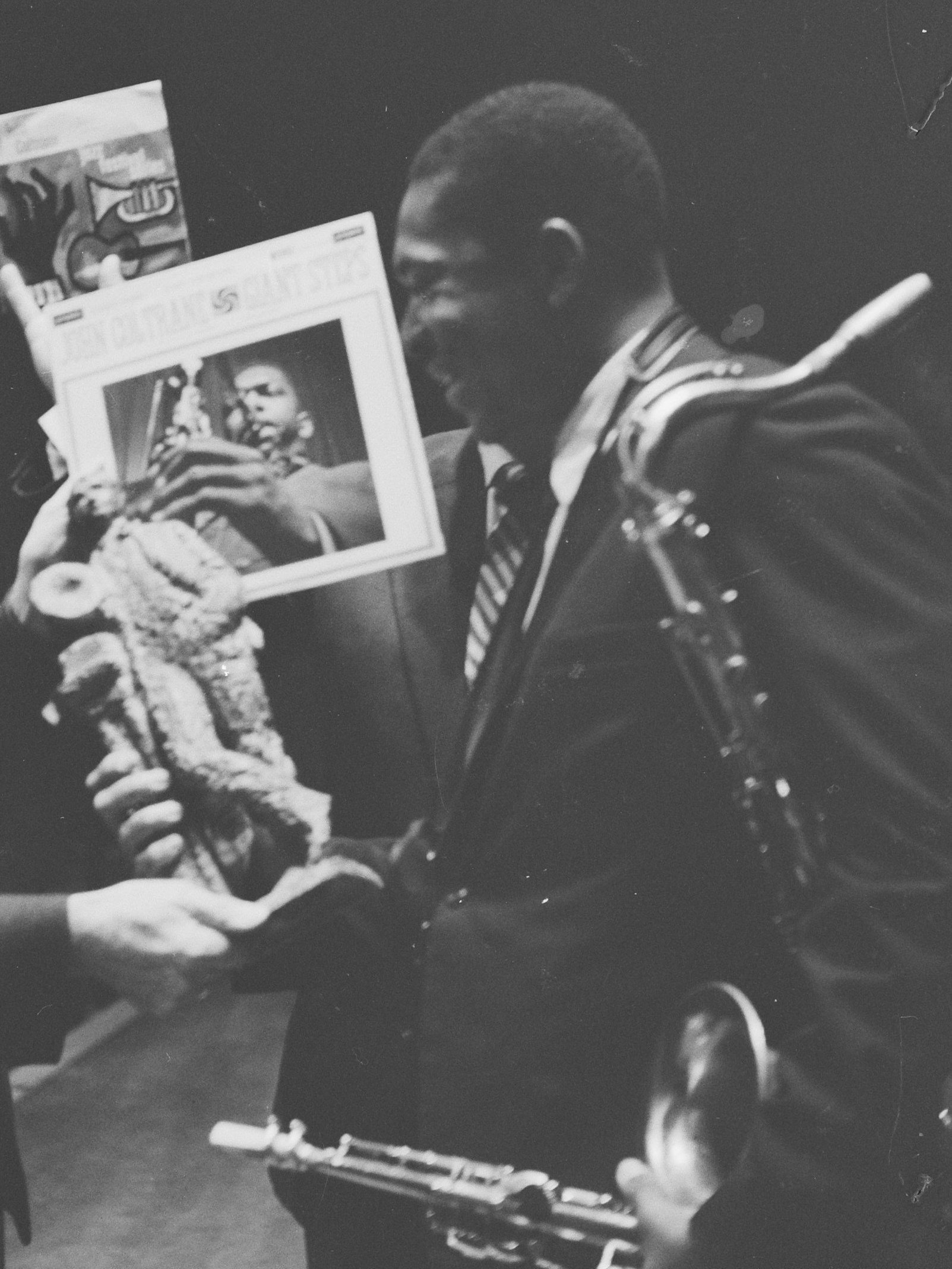 Man in a suit holding a saxophone and a record album, smiling, in a black-and-white photo.