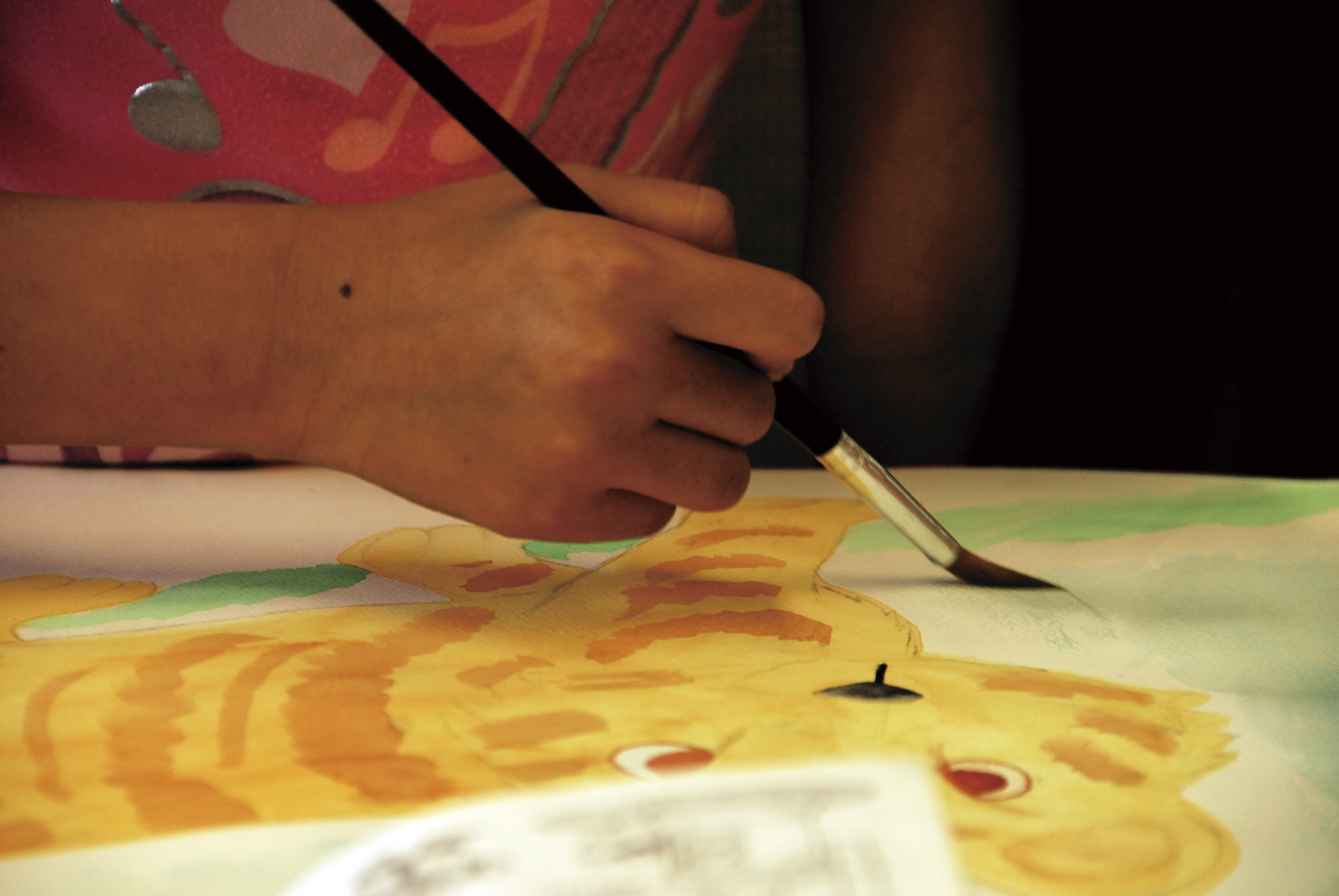 A child's hand holds a thin paintbrush over a colorful watercolor painting.
