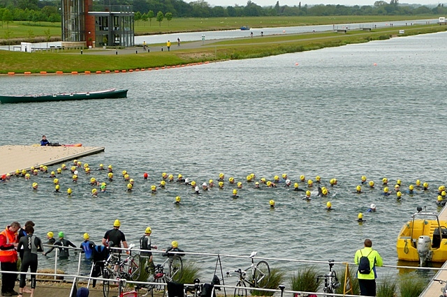 Swimmers in yellow caps wading into open water at the start of a triathlon.