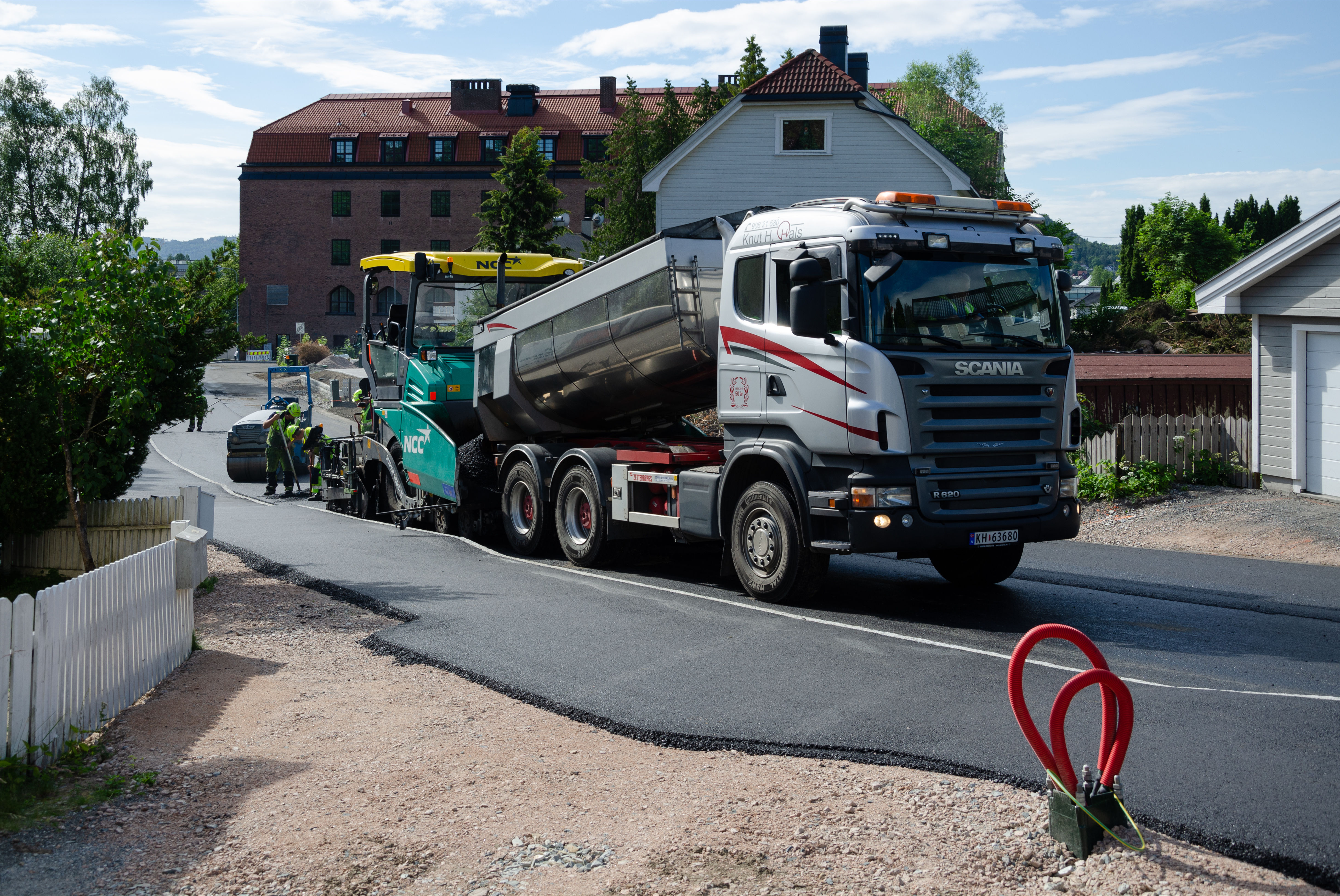 Dump truck and asphalt paver resurfacing a curved residential street with workers in high-visibility vests.