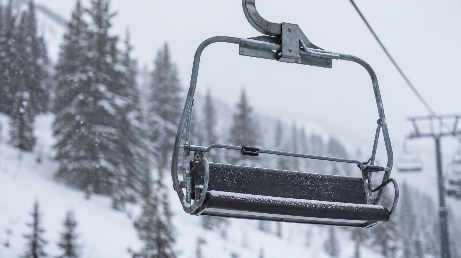 Empty ski chairlift chair on a cable above a snowy slope lined with evergreen trees.