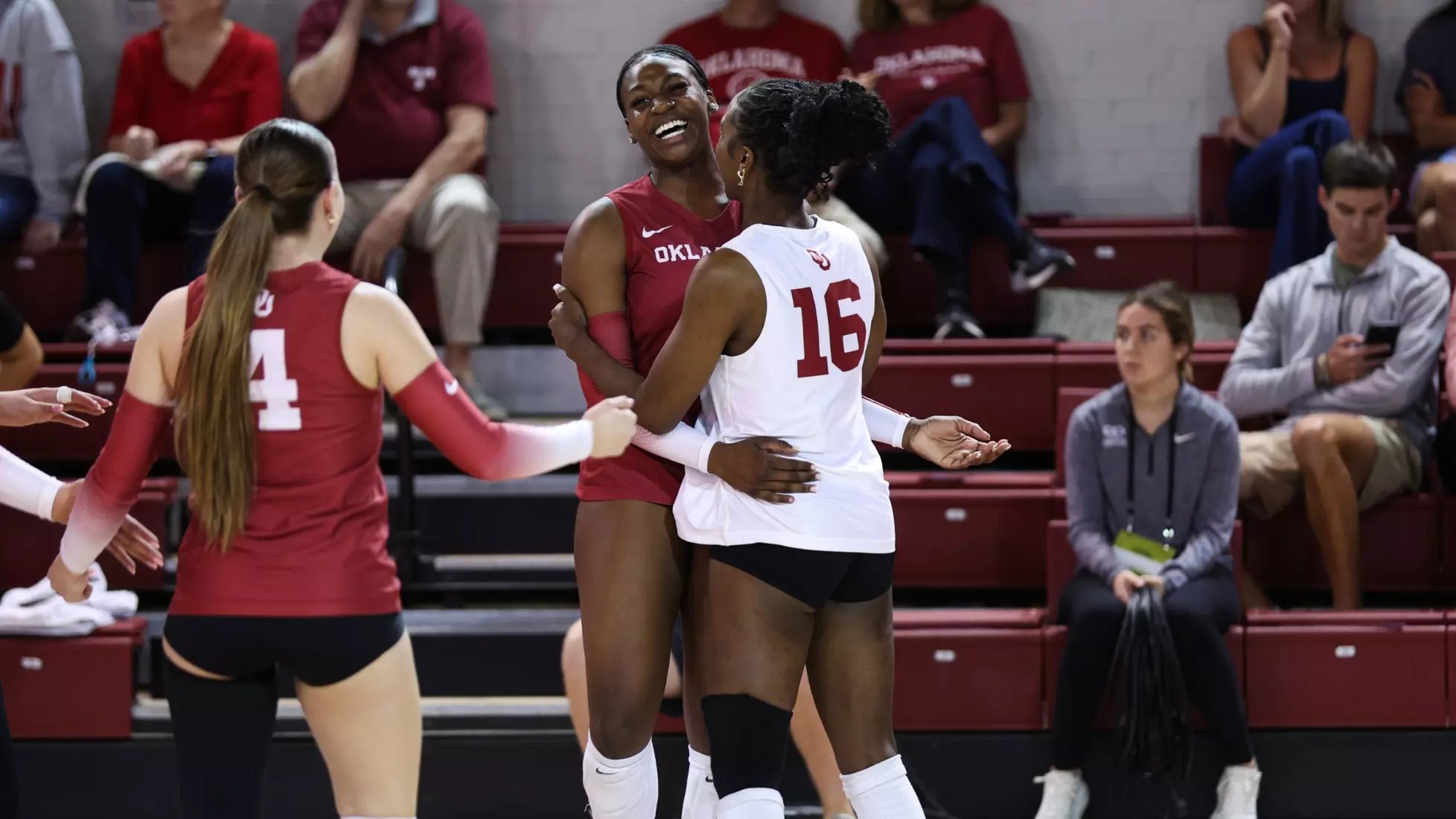Two volleyball players in crimson uniforms embrace and celebrate on an indoor court.