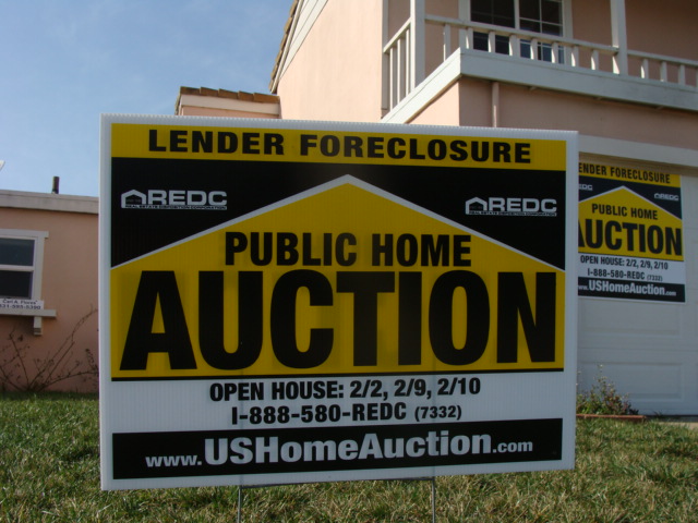 Yellow and black yard sign advertising a lender foreclosure public home auction in front of a house.